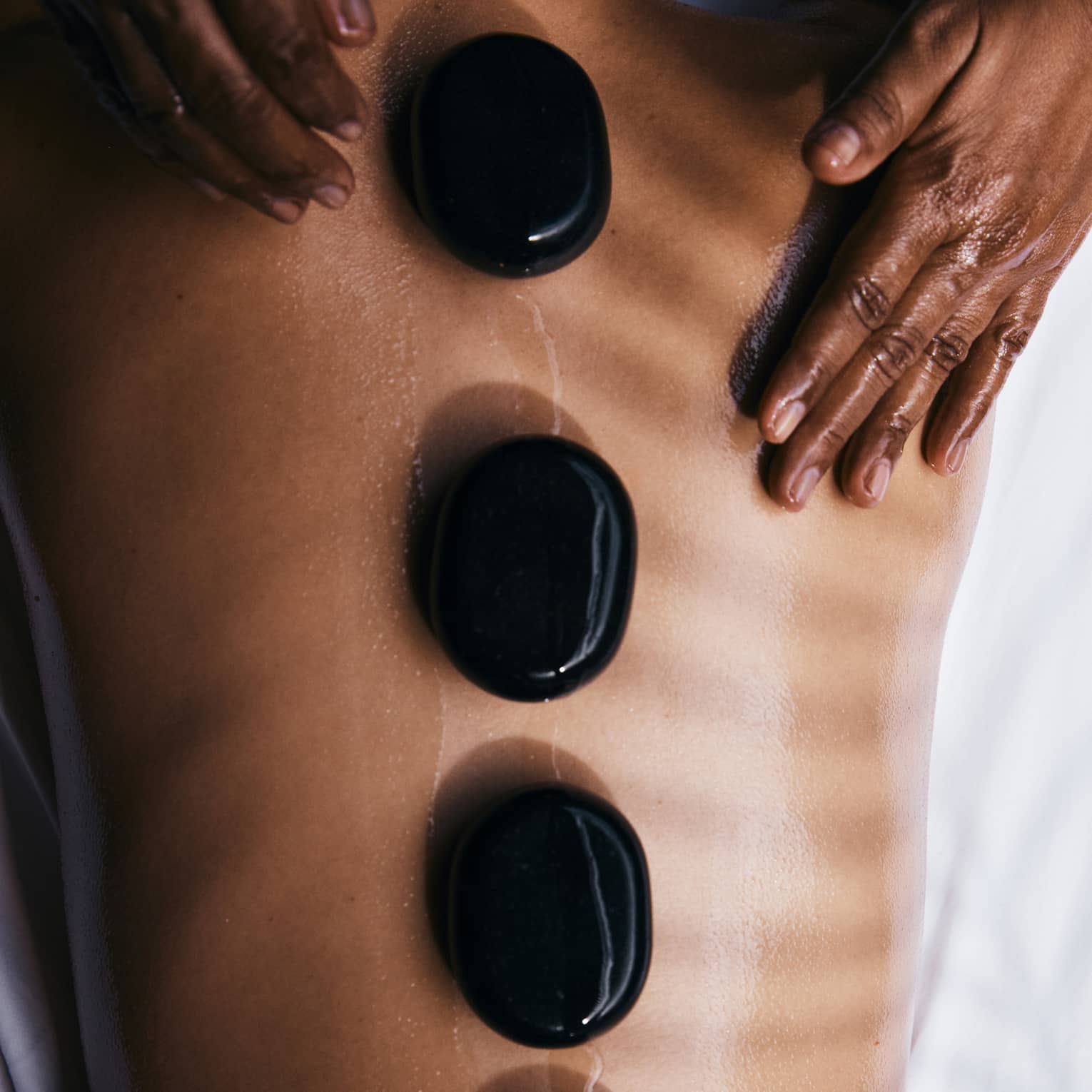 A person lying prone on a spa treatment table with smooth, round, black oval stones placed along their spine