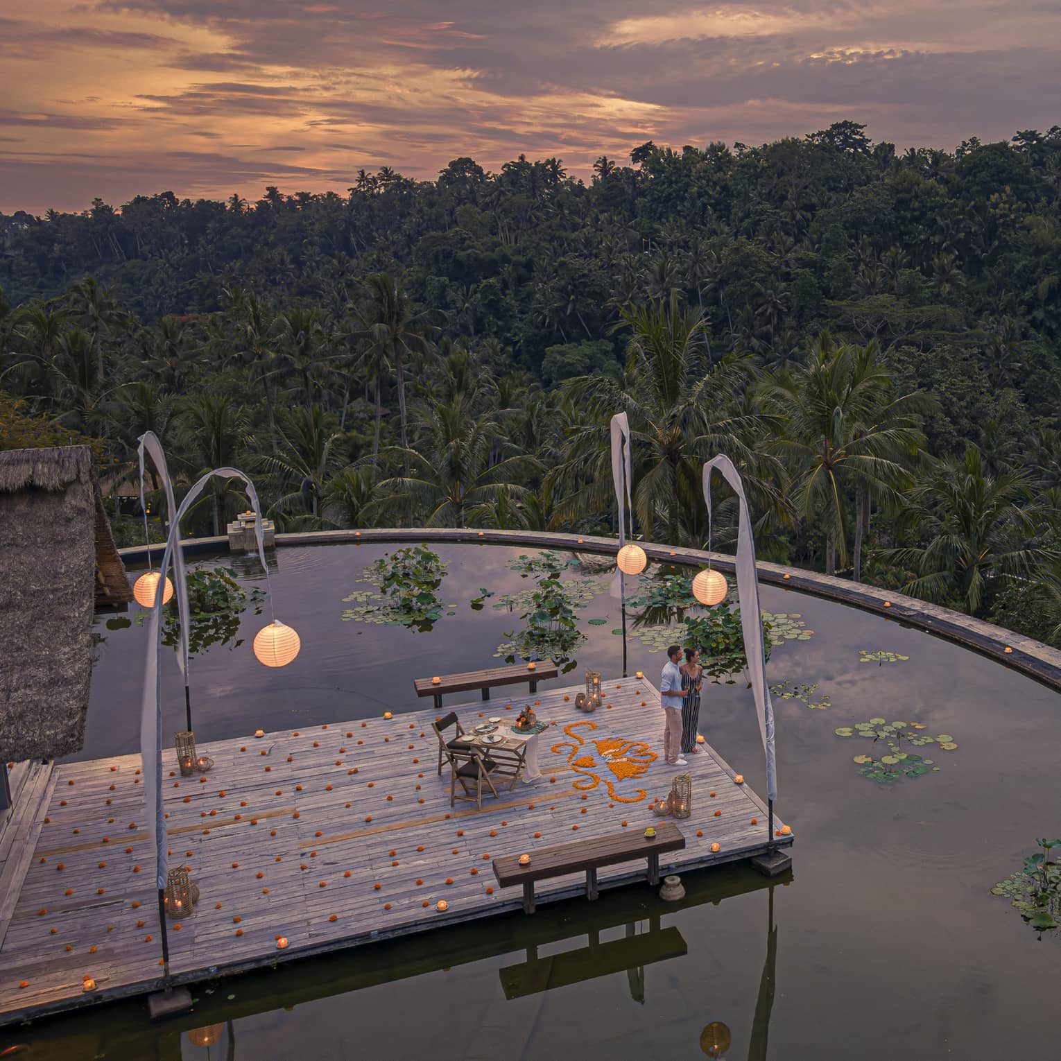 Couple stands on dock at the infinity edge lotus pond overlooking dense forest at dusk