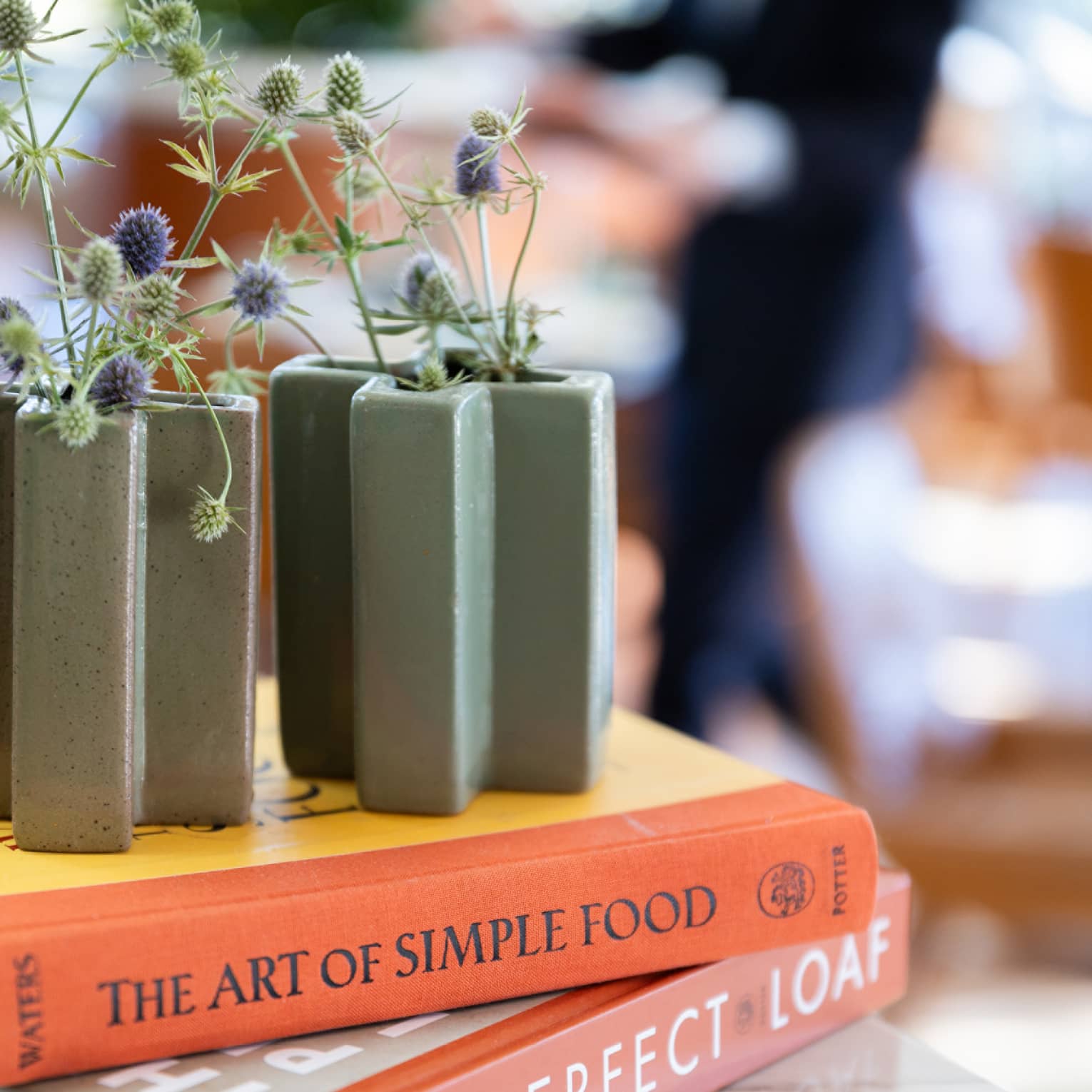 Purple and green flowers in slate-green vases sit on top of two books stacked on a marble table