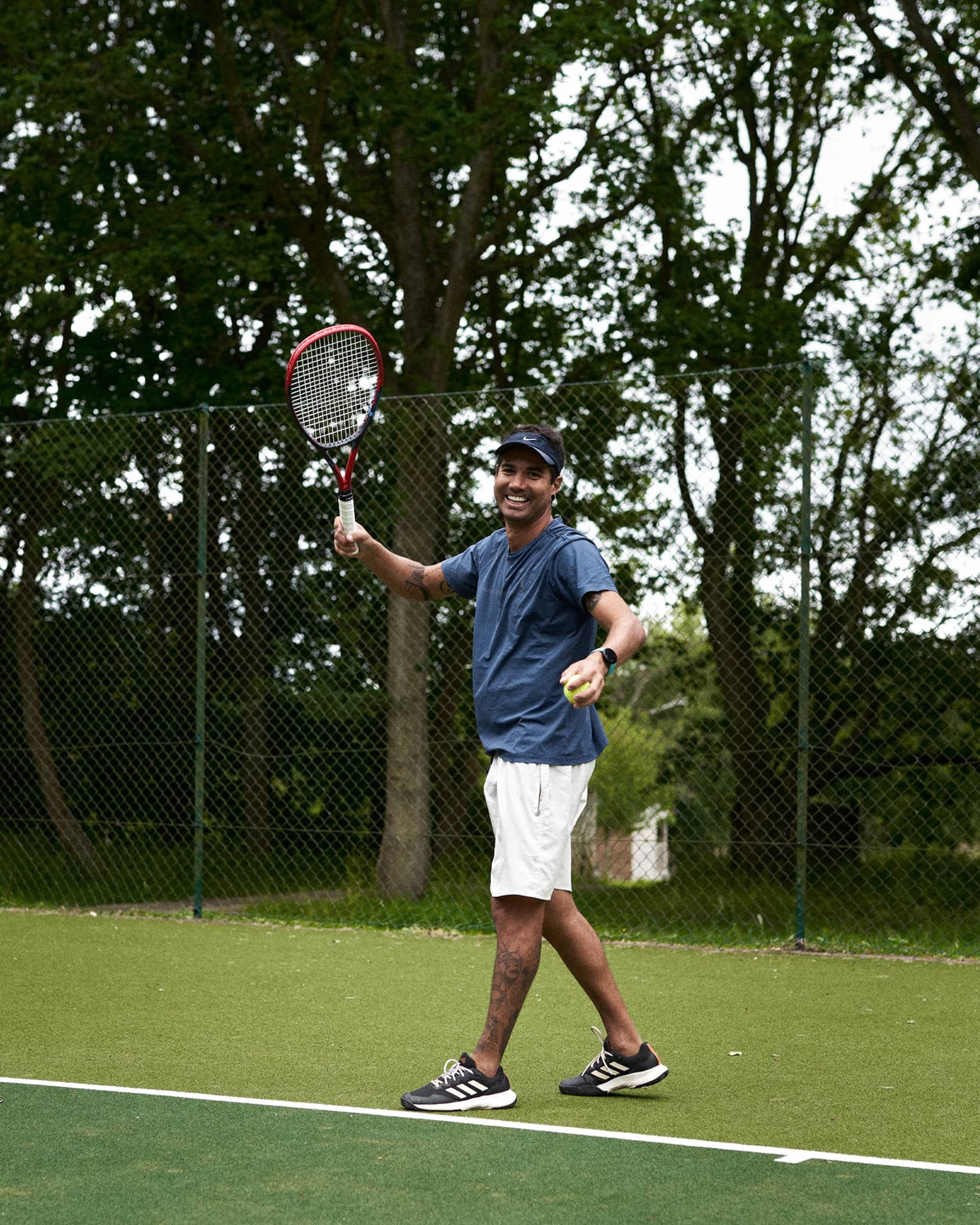 Person standing and smiling while readying to serve on green tennis court with fence and trees in backdrop