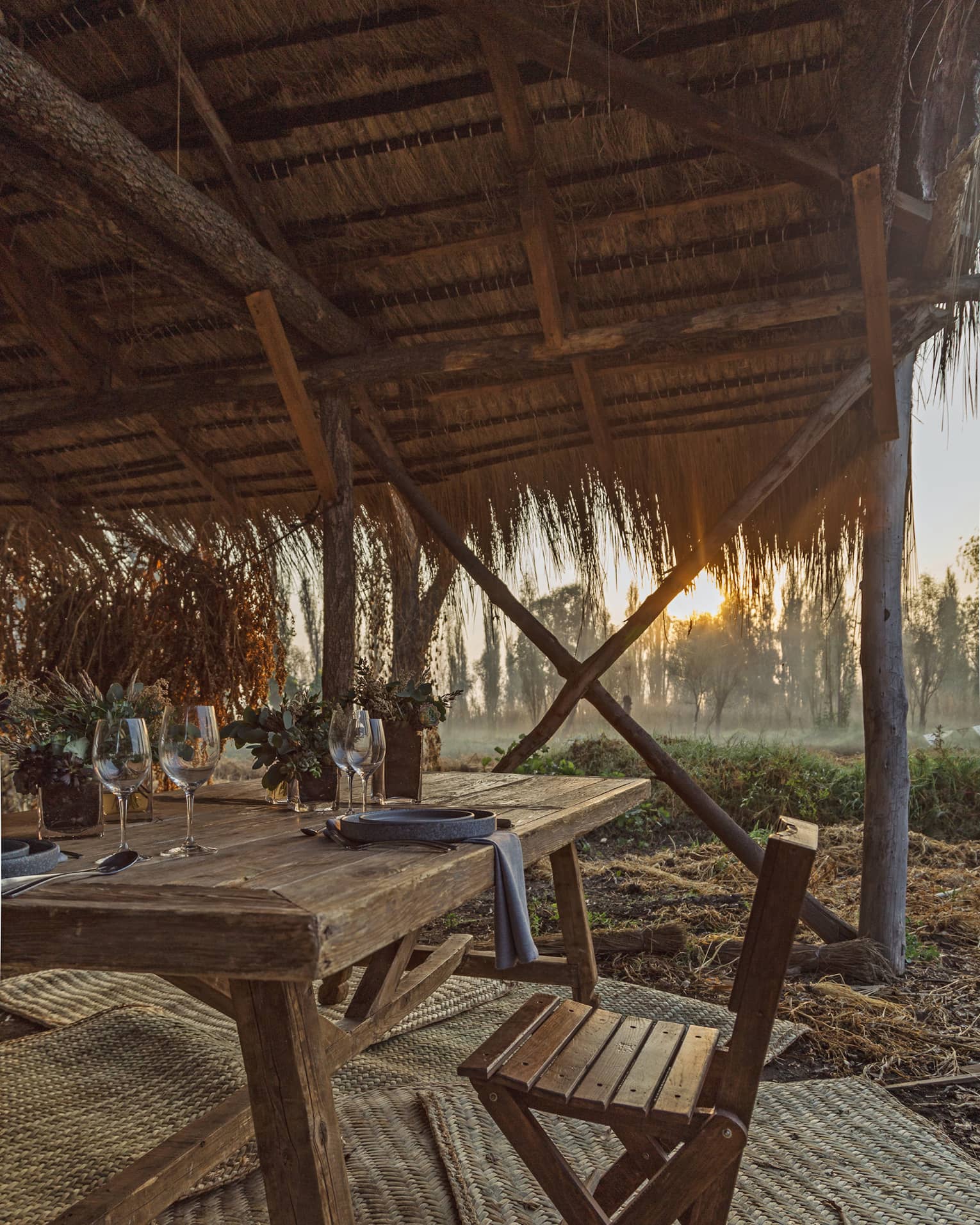 The setting sun shining on a thatched wooden canopy covering a wooden table with rustic dinnerware and succulent plants.