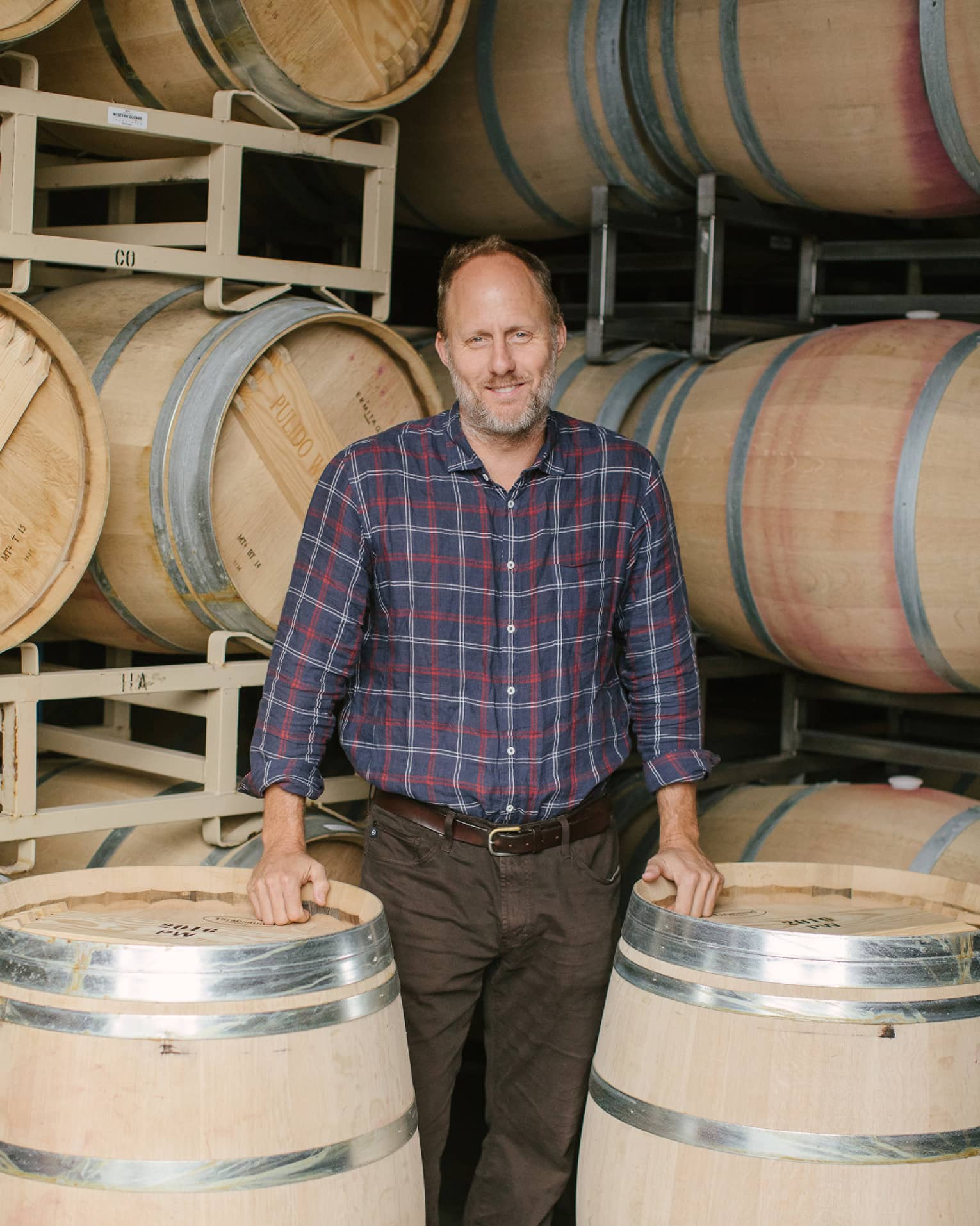 Thomas Brown stands before two racks of wine barrels as he rests his hands on two upright barrels and smiles at the camera.