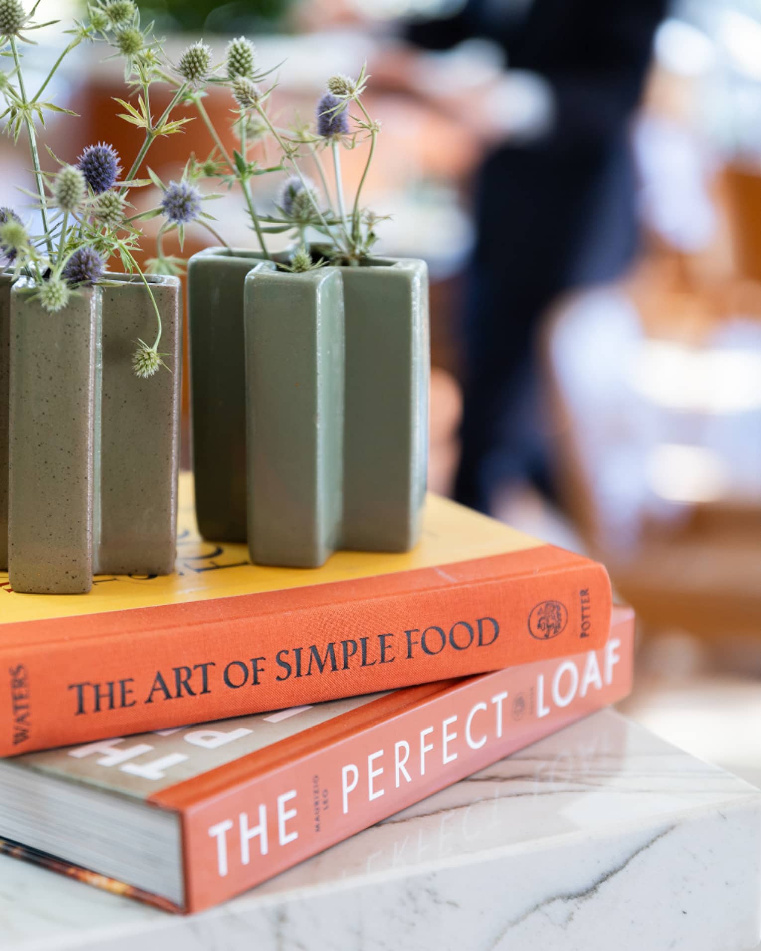 Purple and green flowers in slate-green vases sit on top of two books stacked on a marble table