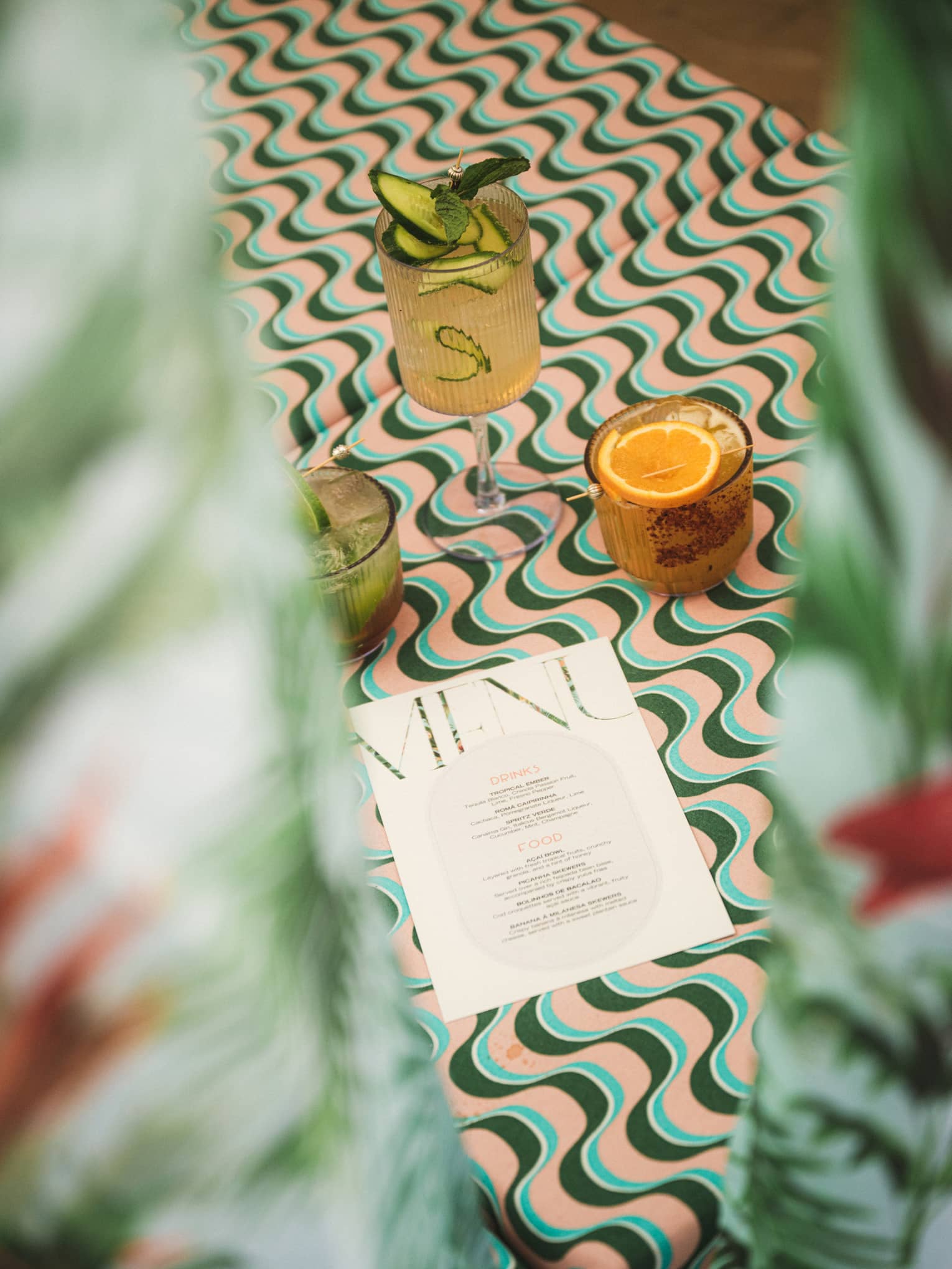 A paper menu lies on a pink-blue-and-green-wave patterned daybed next to three cocktails