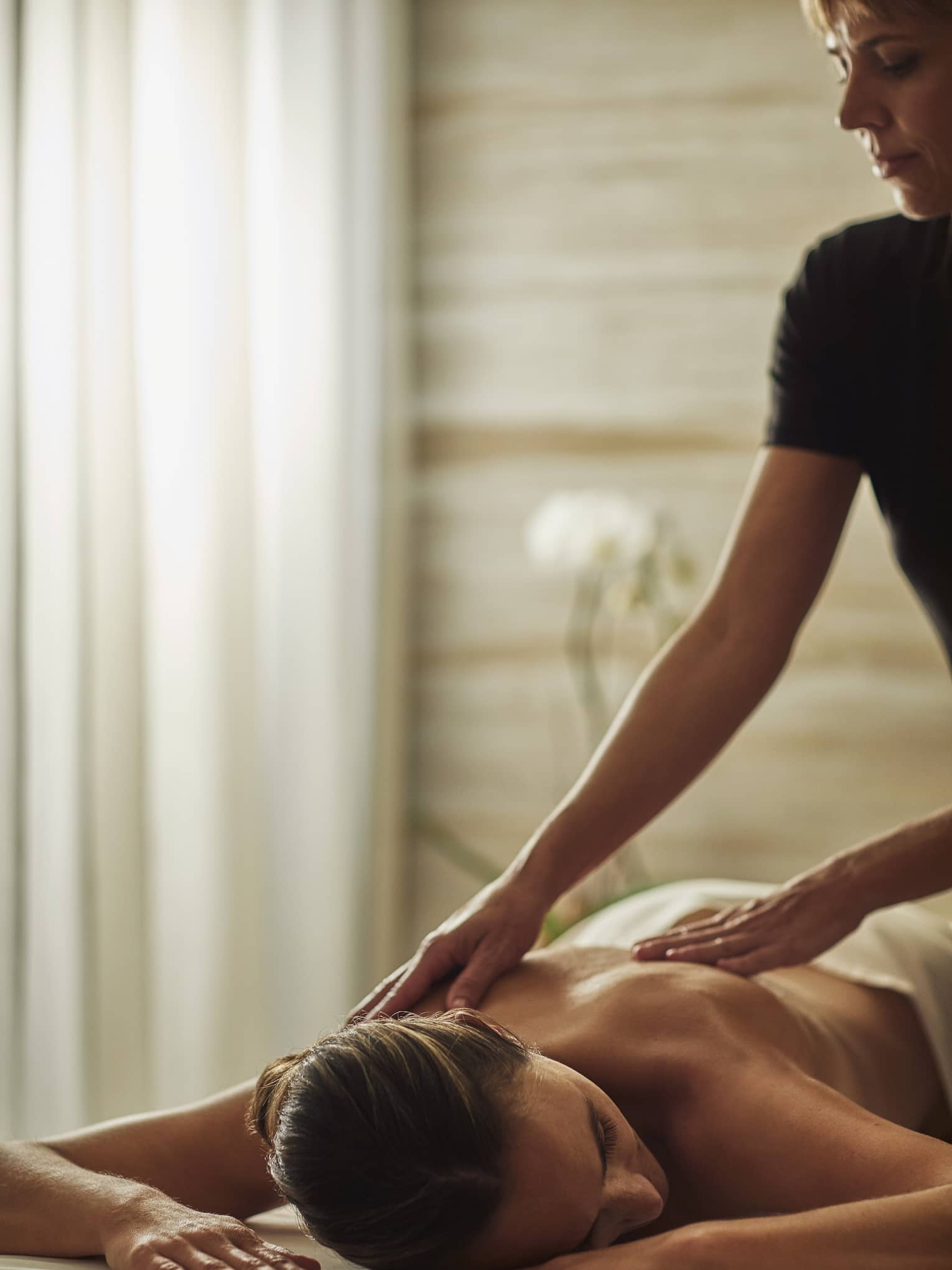 Woman in black uniform massages bare shoulders of woman lying on table face-down with head to side