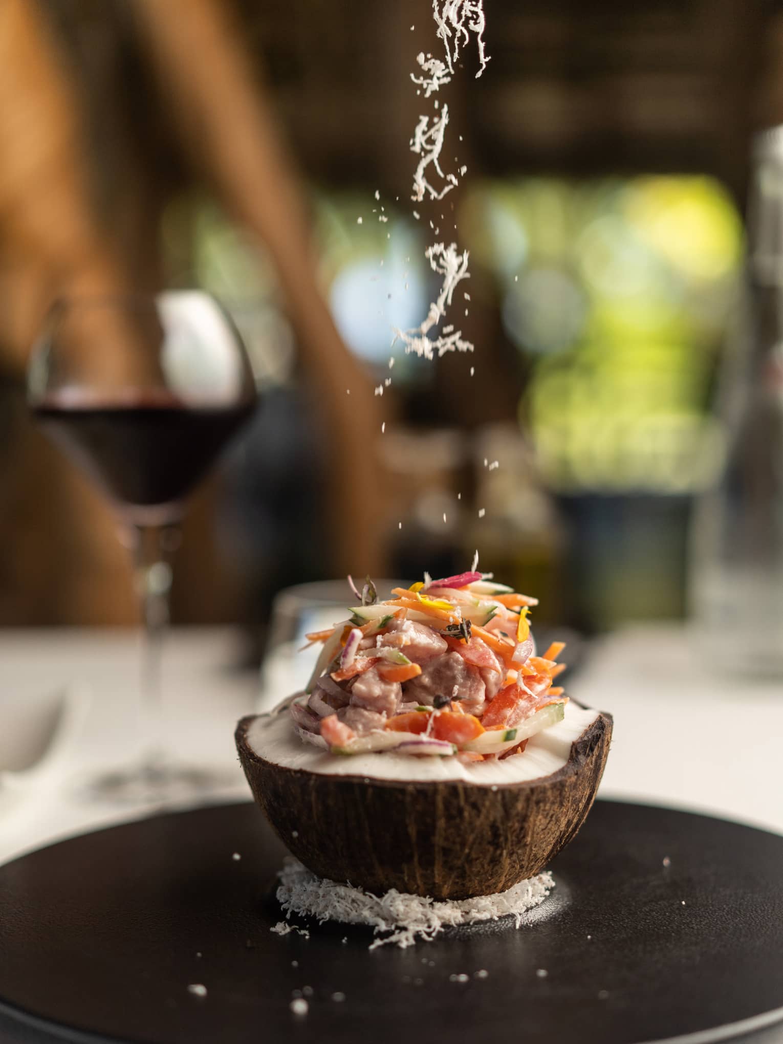 Close-up of an open coconut filled with fish and thinly sliced vegetables, atop a bed of coconut flakes on a round platter.