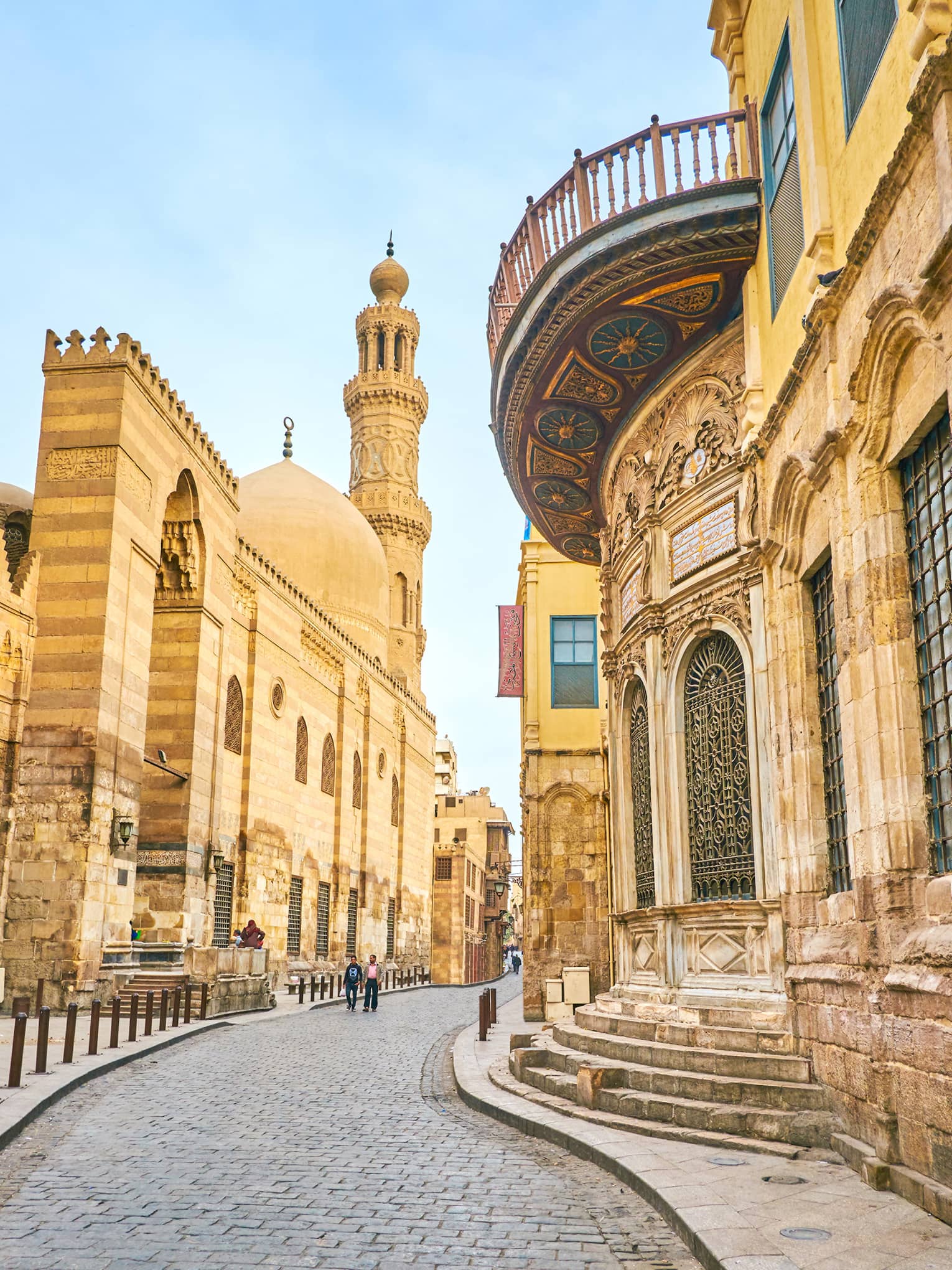 Pedestrians walk along a meandering cobbled road between tall, yellow stone buildings with ornate lattice windows.