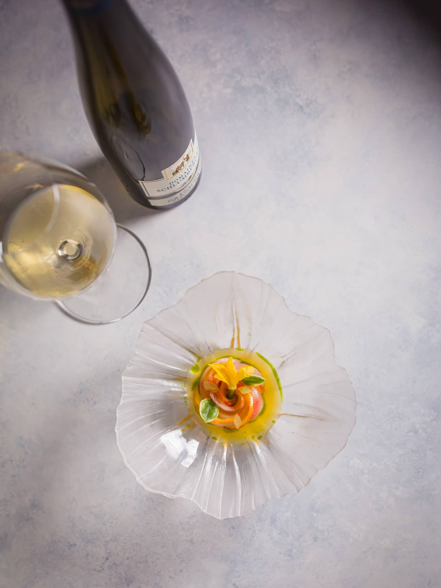 Overhead view of a colourful appetizer garnished with florals in a glass, flower-shaped dish beside a glass of white wine.