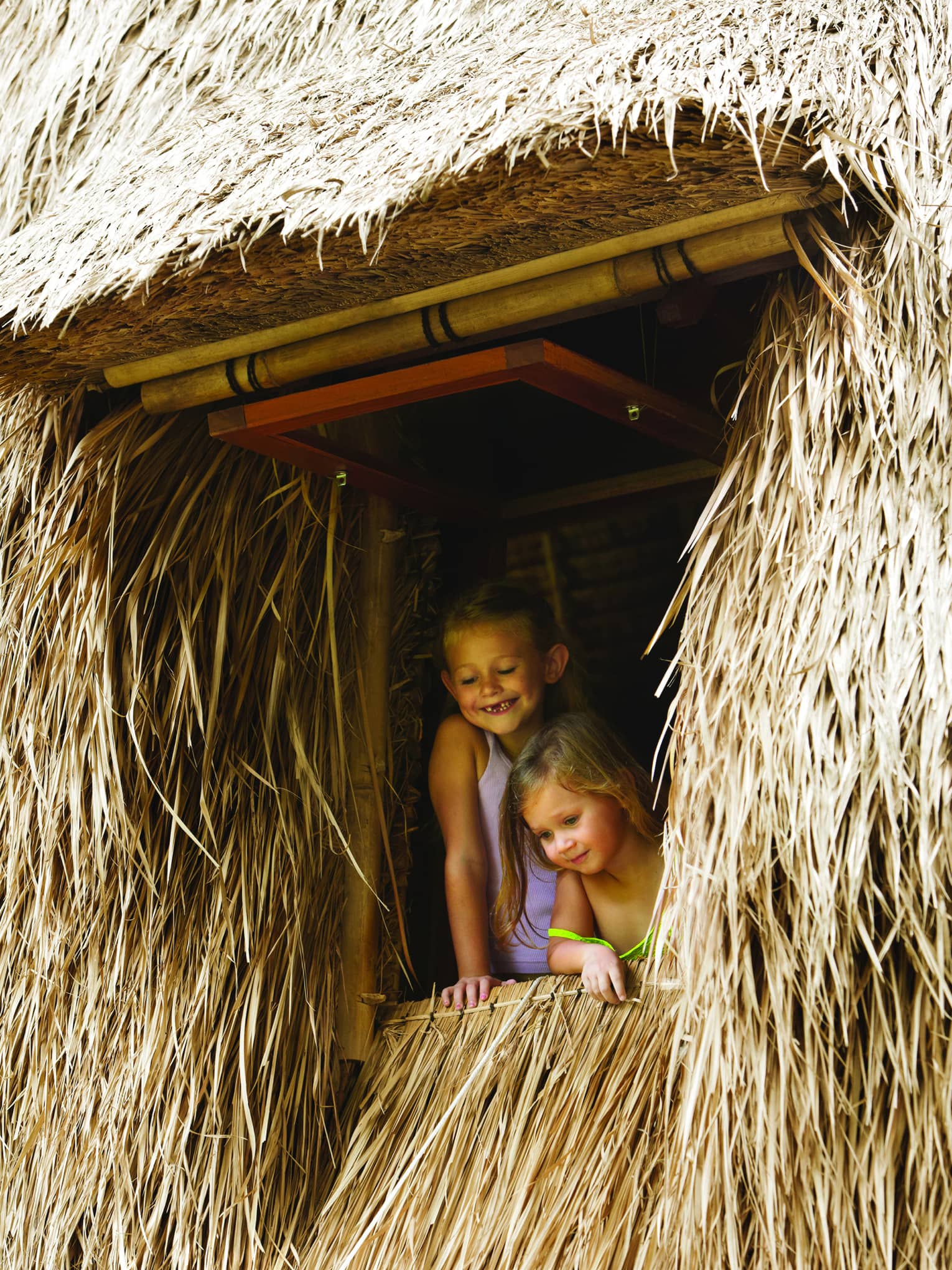 Two smiling young children look out from window of thatched building