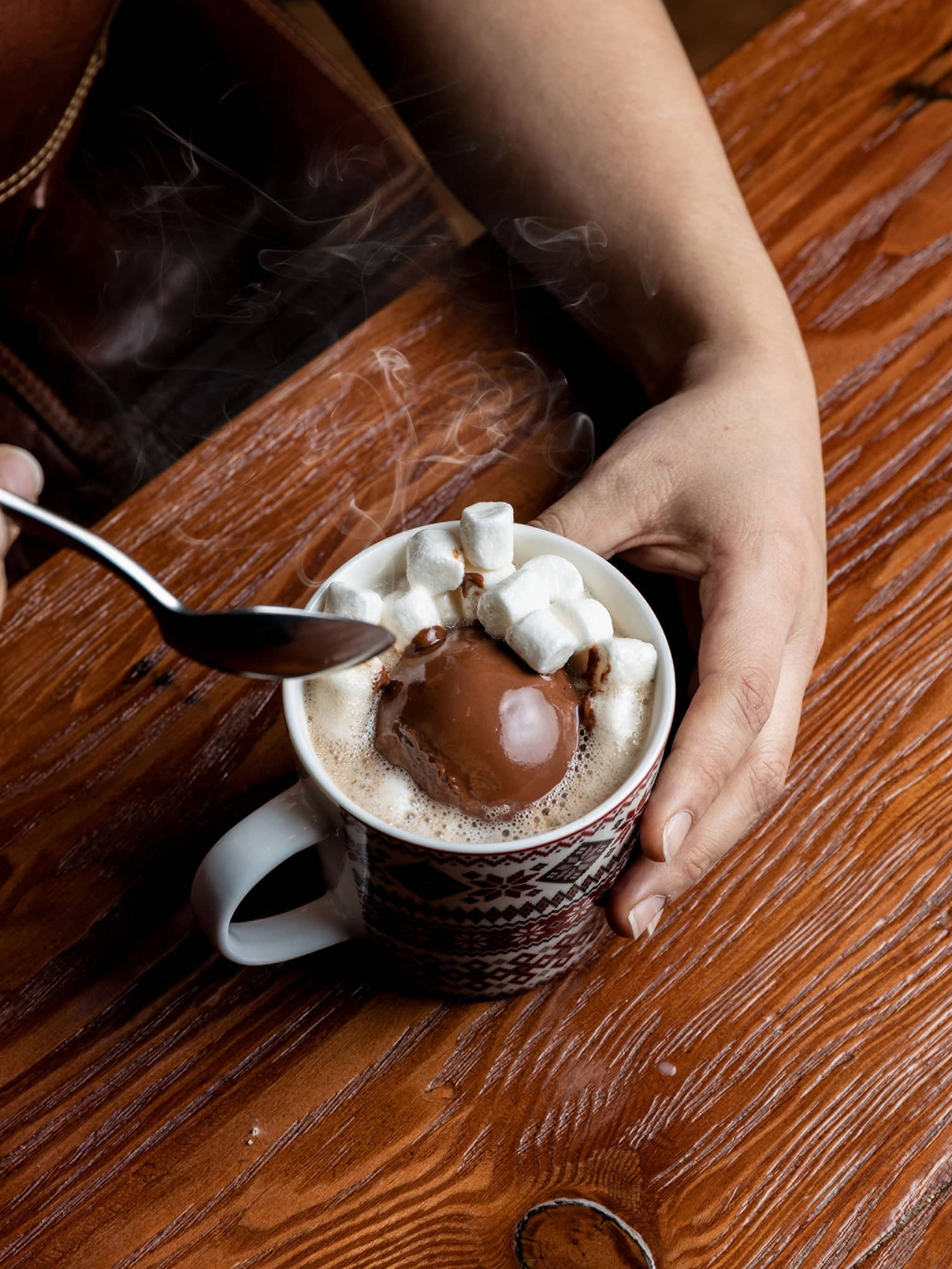 Natural wood table with a hand curled around a mug filled with steaming hot chocolate topped with numerous small marshmallows, and the other hand holding a spoon