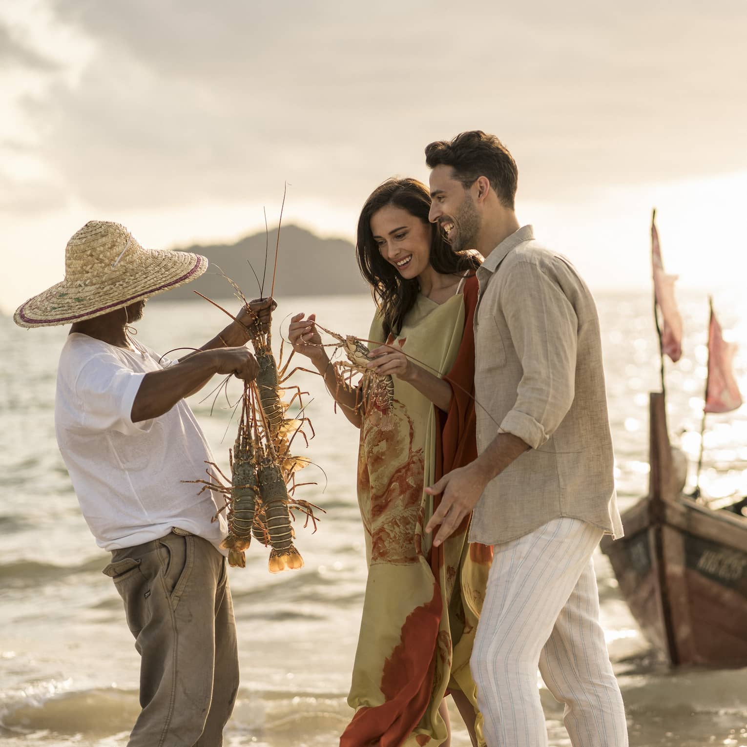 Local fisherman holds up two freshly caught lobsters for couple, traditional fishing boat in background at sunset