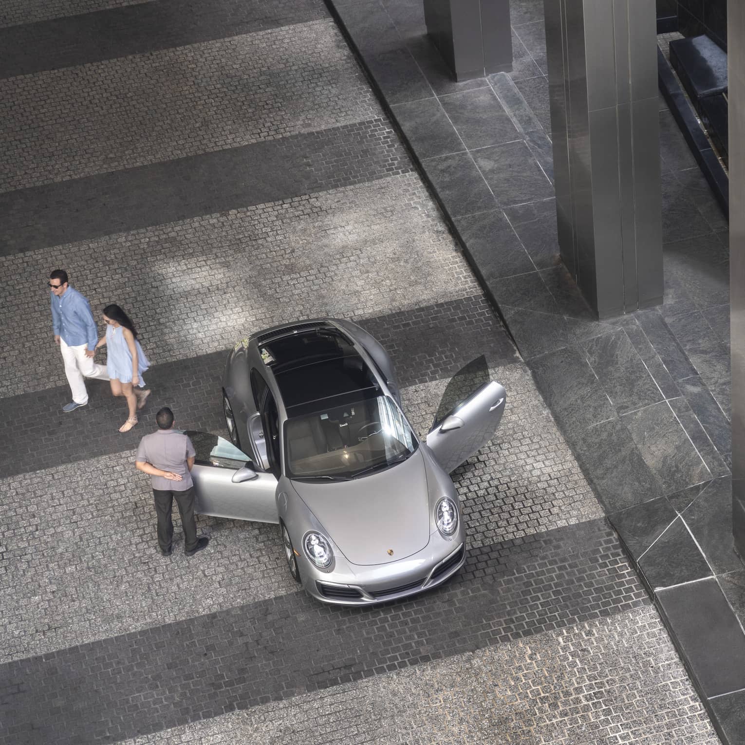 Aerial view of hotel staff opening door of luxury silver car on driveway, couple walking in background