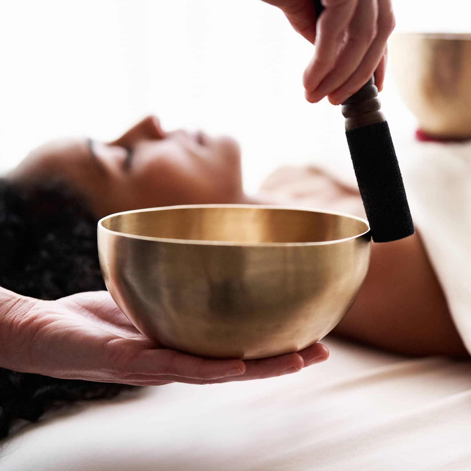 Close up of hands playing a singing bowl with a woman laying down on a spa treatment table in the background