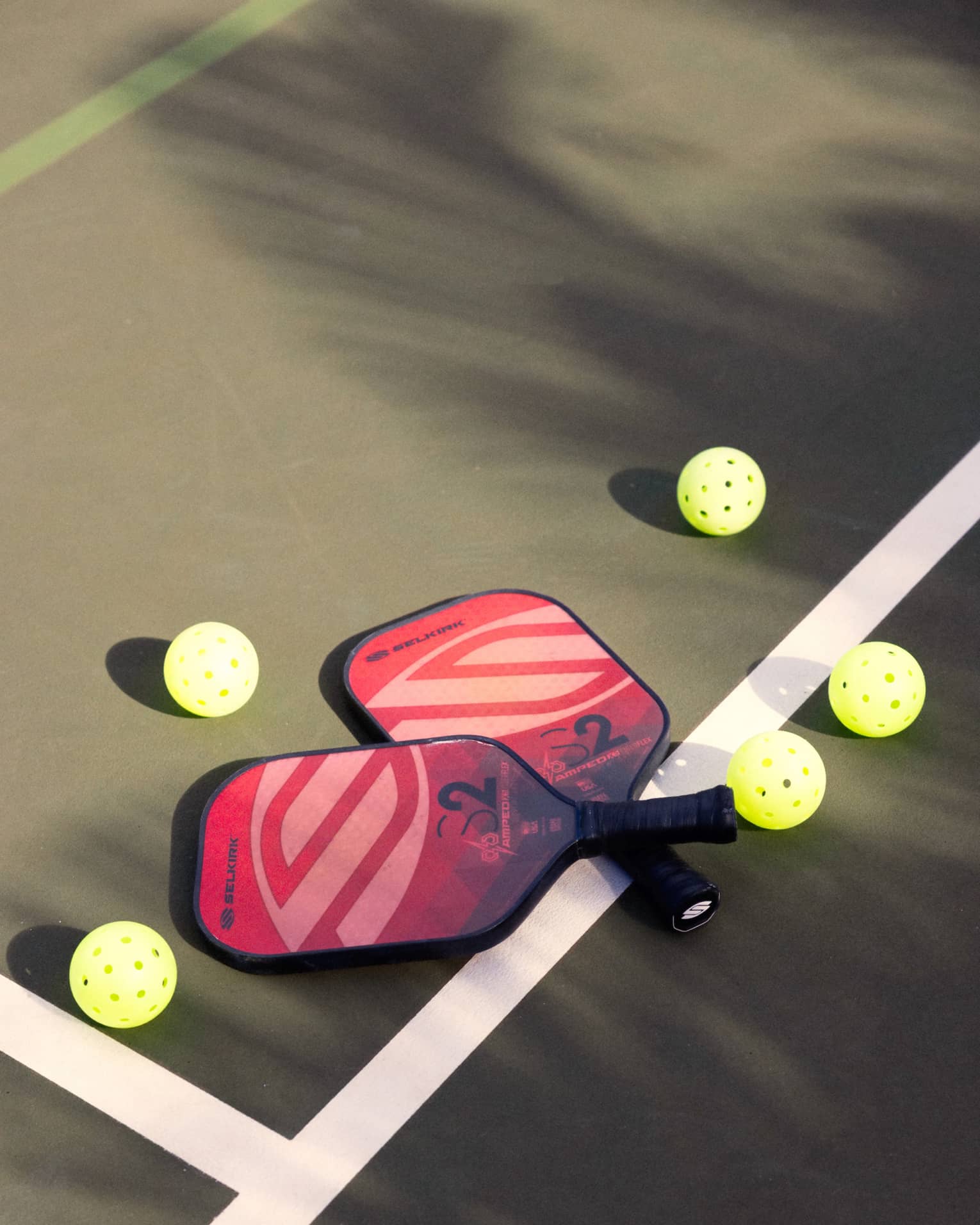 Close-up of two red and pink pickleball racquets, handles crisscrossed on a white court line, surrounded by five pickleballs.