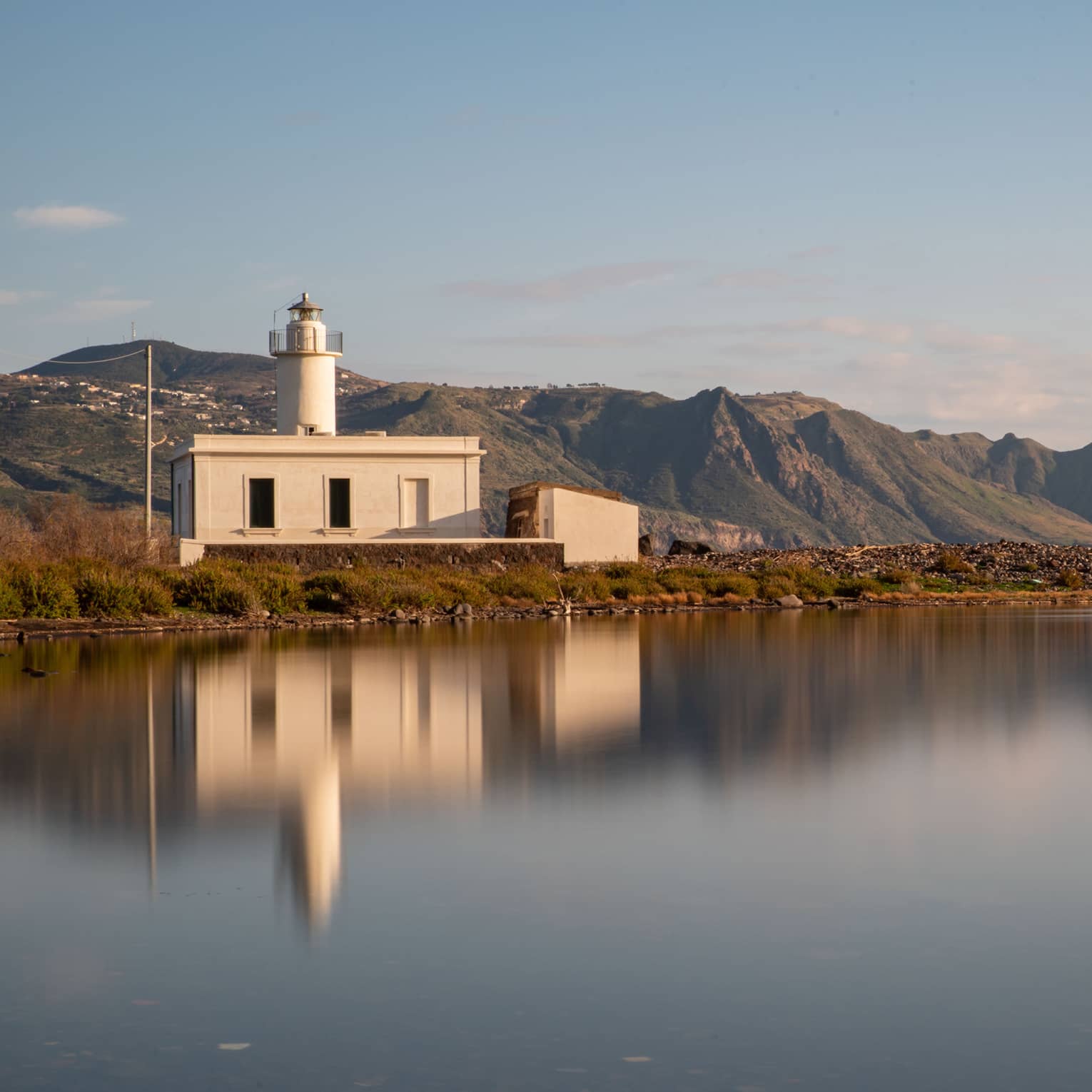 Short all-white lighthouse, reflecting on smooth water, mountains and blue sky in backdrop