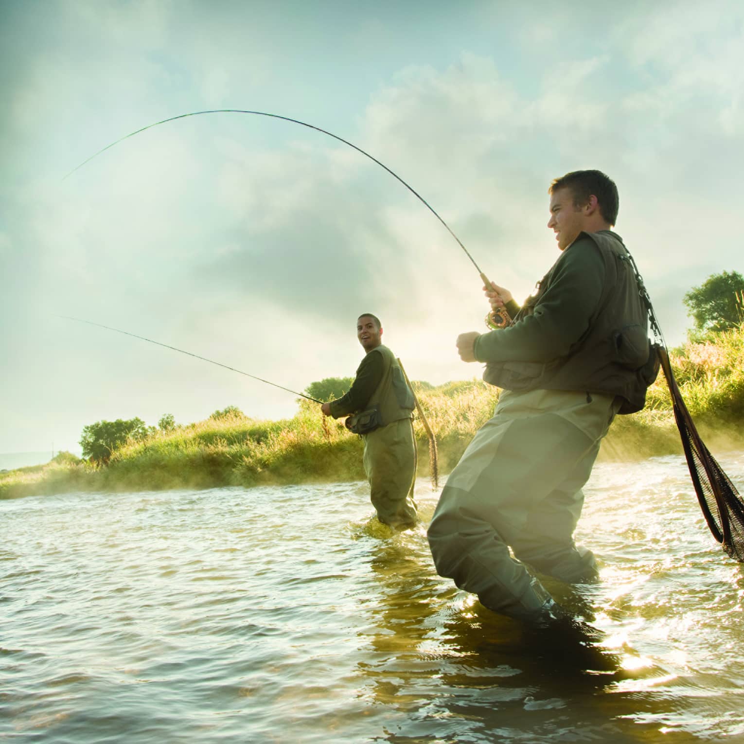 Two people fly fishing in a river, standing knee-deep in the water, surrounded by mist and greenery.