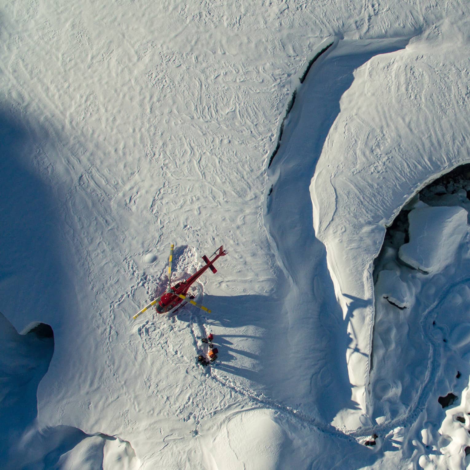 Aerial view of a red helicopter that's landed on a snow-covered mountain with three people walking near it