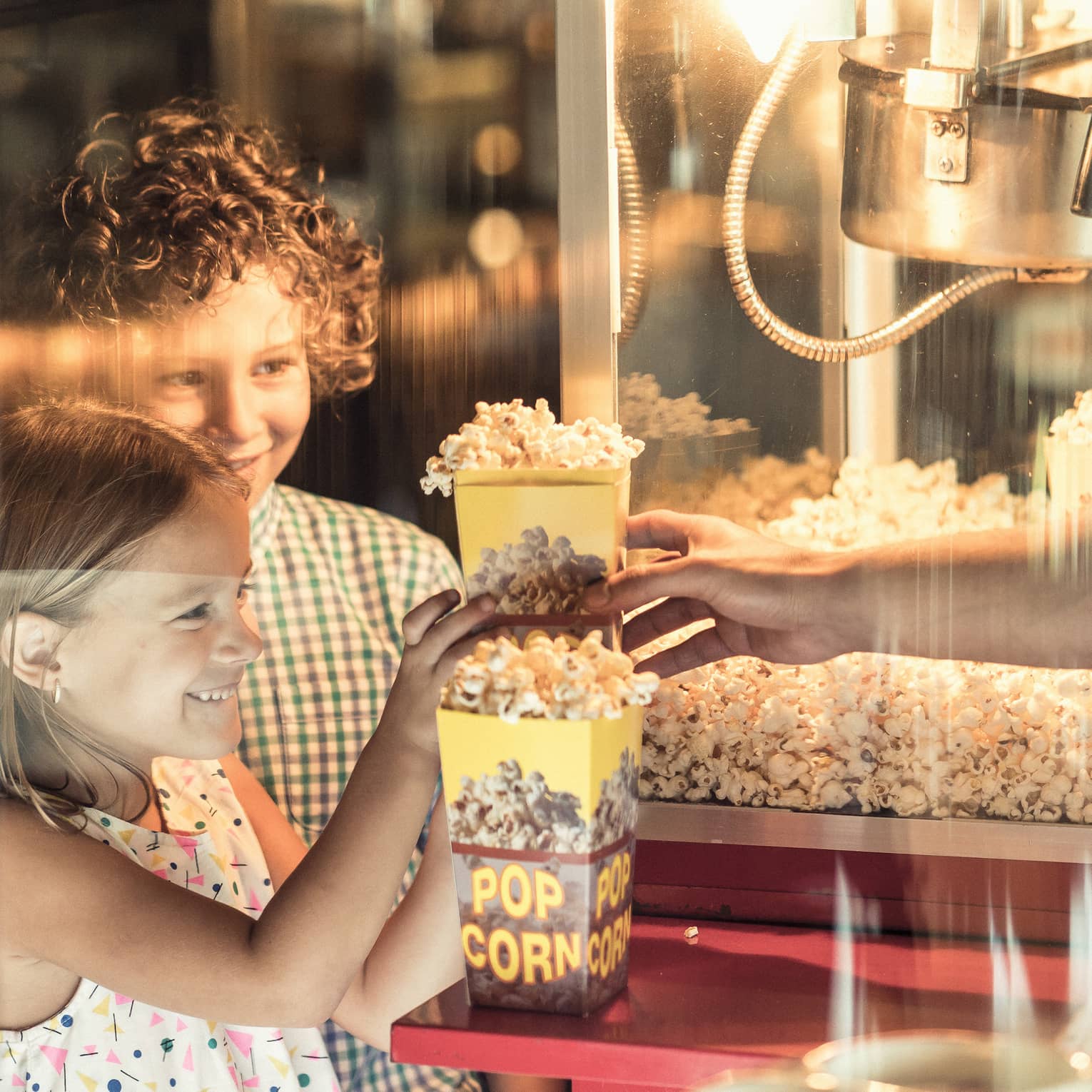 Two children smile in the warm glow of a movie-style popcorn machine as they receive boxes of popcorn from a vendor.