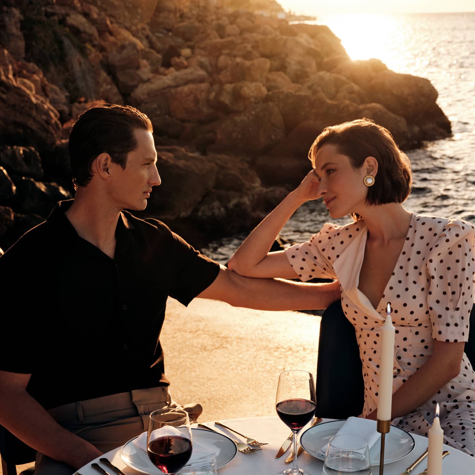 Two people sit at an elegant dining table in an outdoor setting, with a rocky shoreline and the ocean reflecting the sun in the background.
