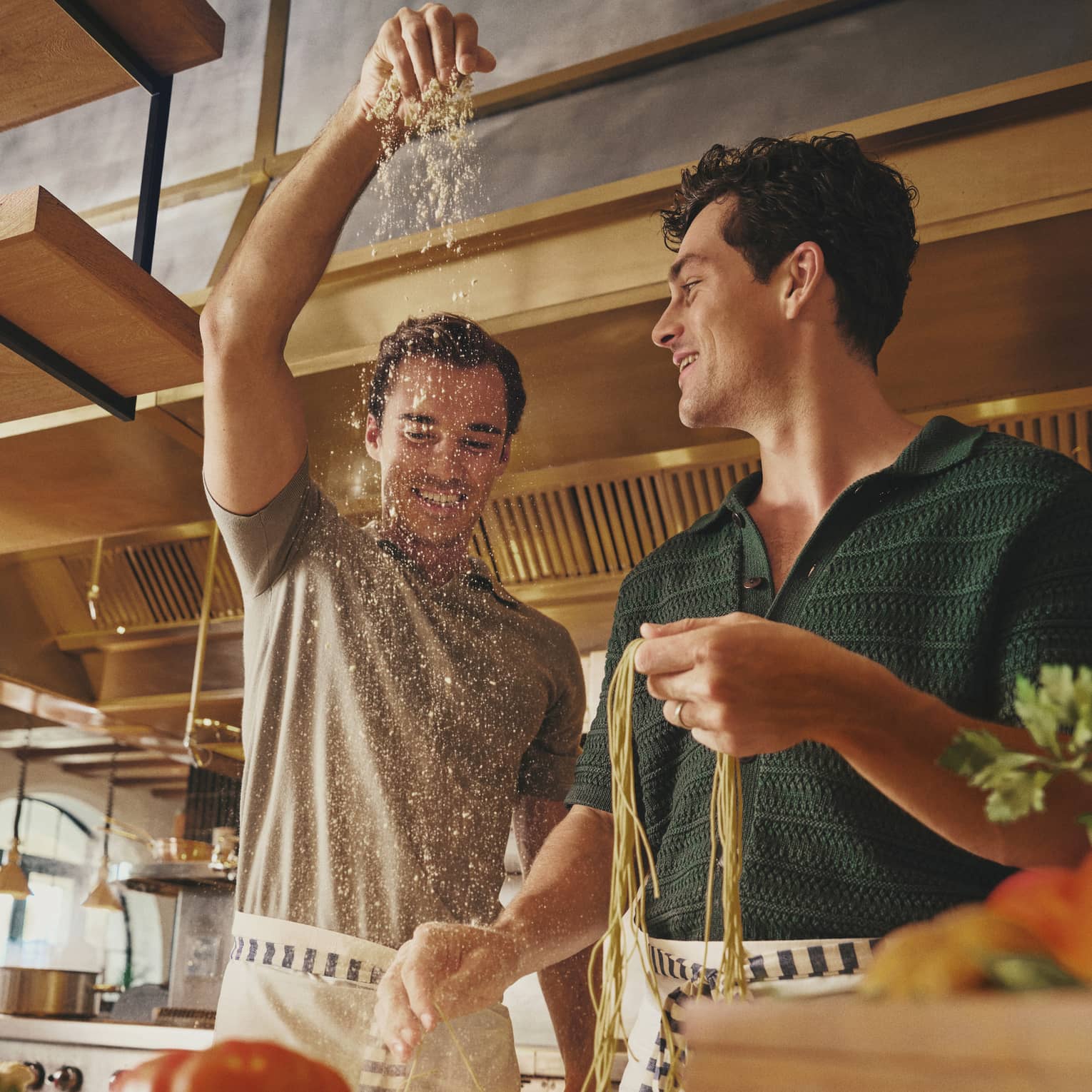 Two smiling guests in aprons prepare fresh pasta noodles on a counter laden with vibrant fresh produce in a bright kitchen.