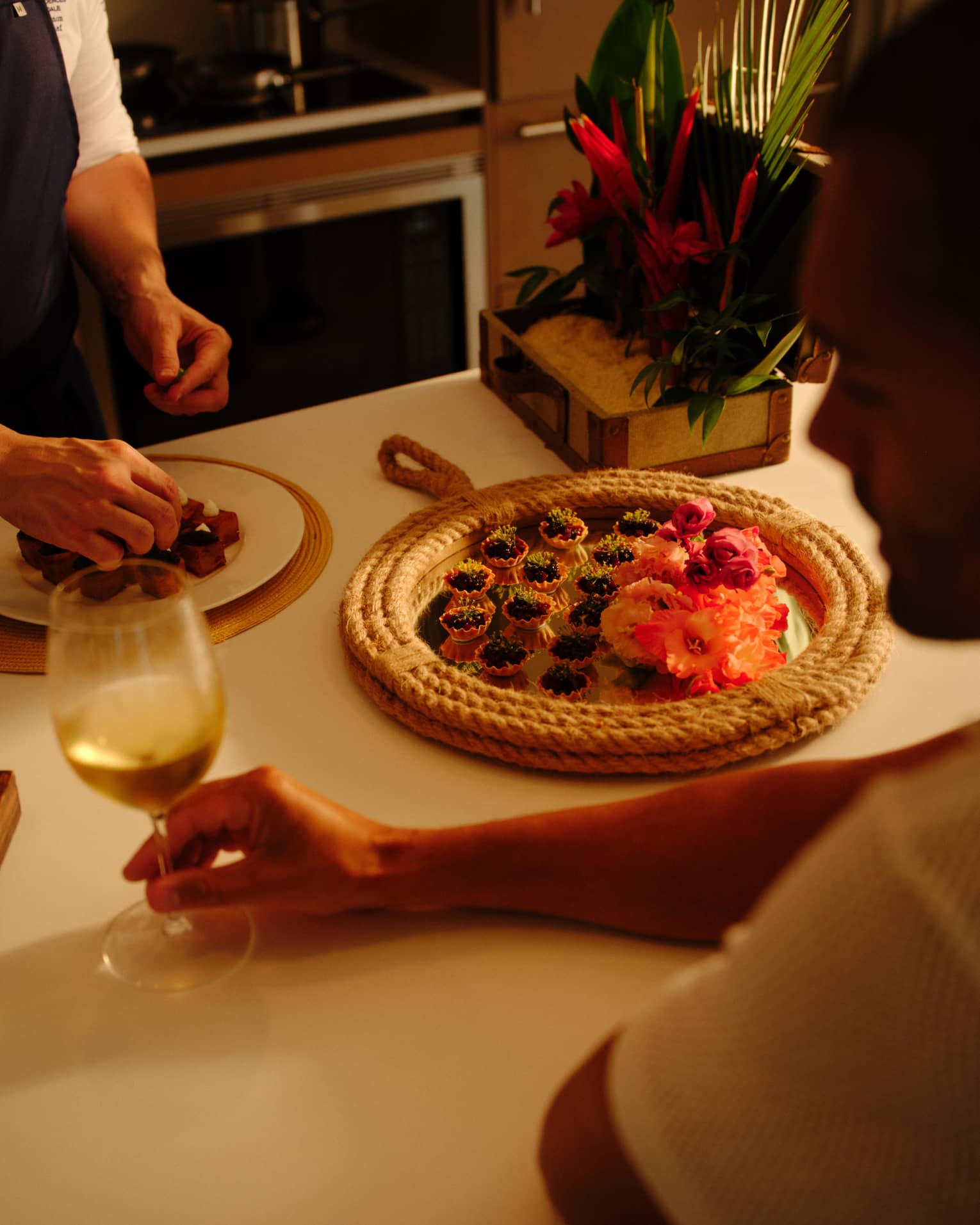 Person sits at a kitchen counter holding a glass of white wine as a chef prepares a plate of appetizers,Person sits at a kitchen counter holding a glass of white wine as a chef prepares a plate of appetizers
