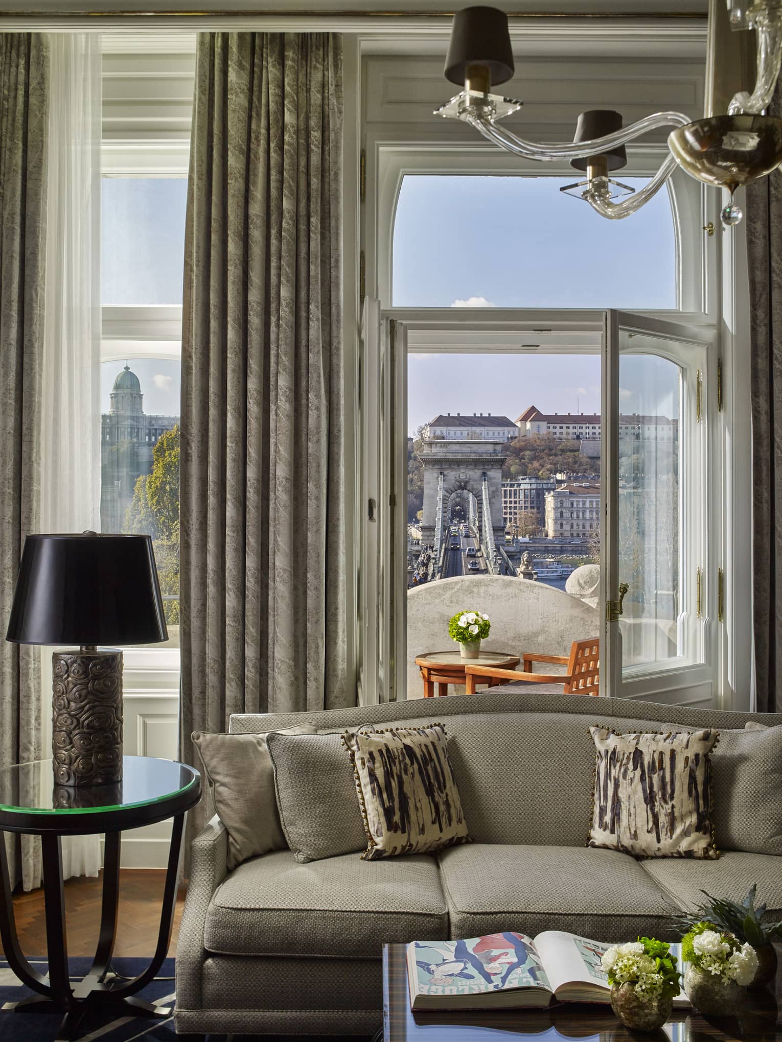 Sofa with patterned pillows facing a balcony with table and chairs overlooking a historic bridge