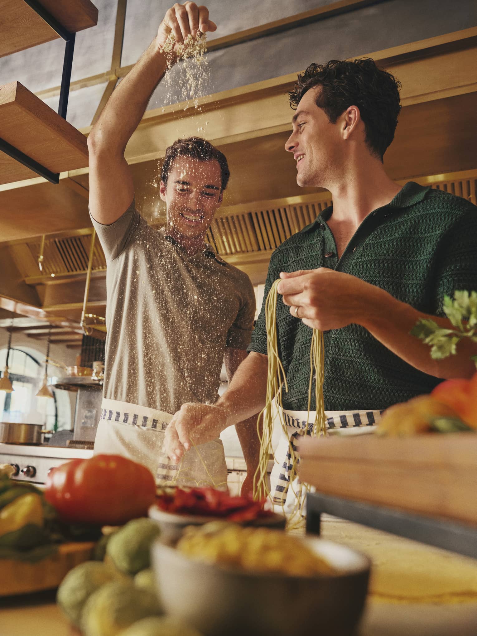 Two smiling guests in aprons prepare fresh pasta noodles on a counter laden with vibrant fresh produce in a bright kitchen.
