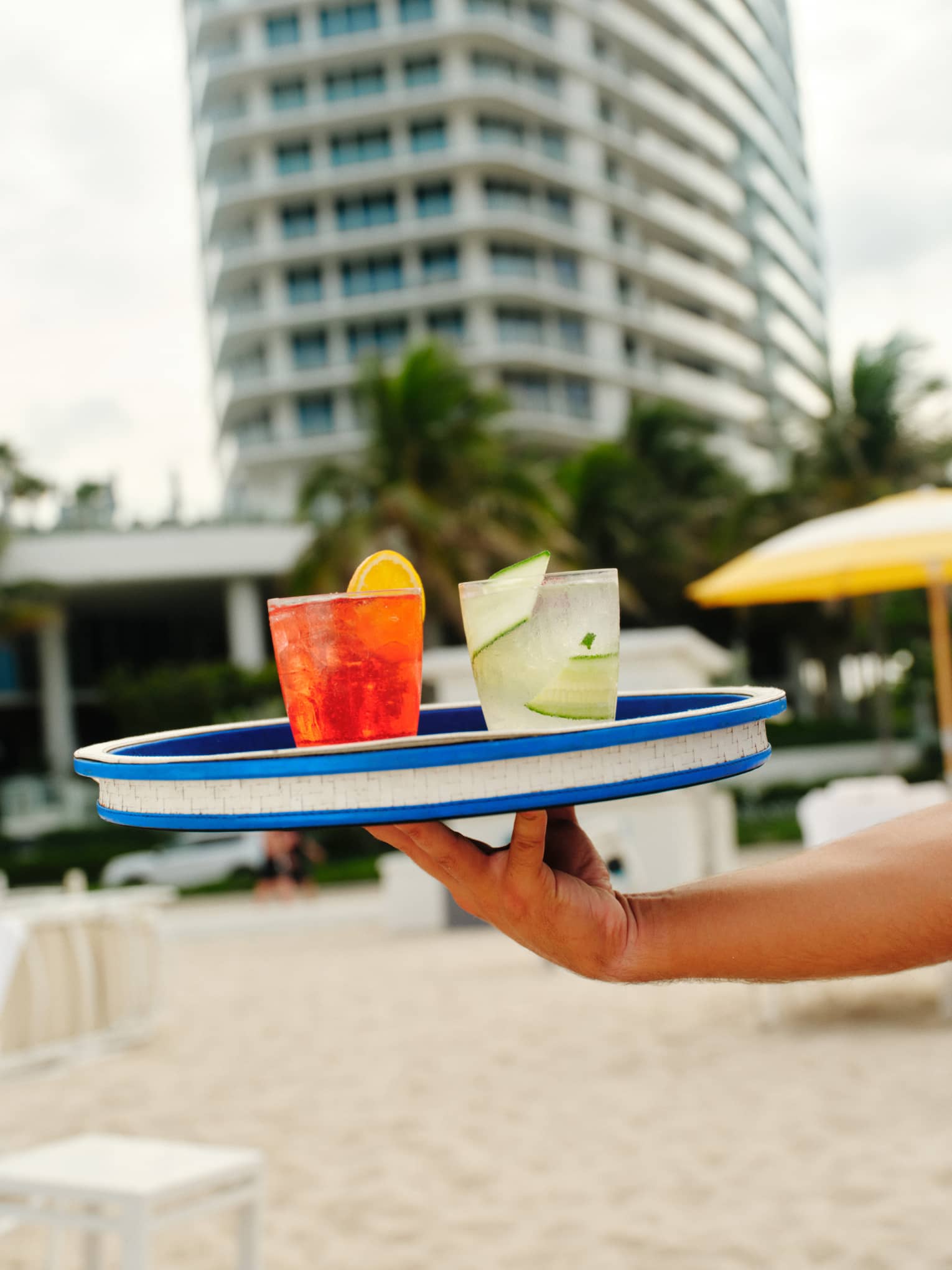 An extended hand holds a blue-and-white serving tray carrying two cocktails to a guest on the beach