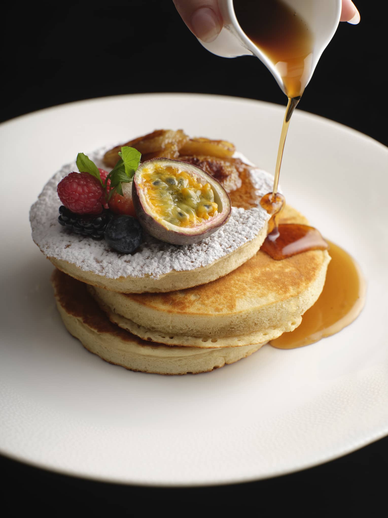 Stack of pancakes topped with powdered sugar and fruit, syrup being poured