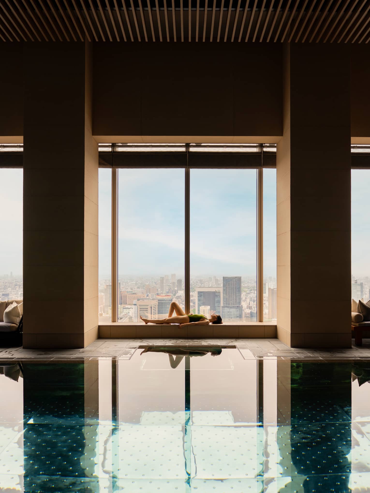 A woman lies at the edge of an indoor pool next to wall of windows overlooking city