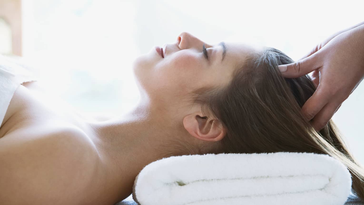 Woman lays on back with head on folded white towel, closes eyes as hands massage her scalp