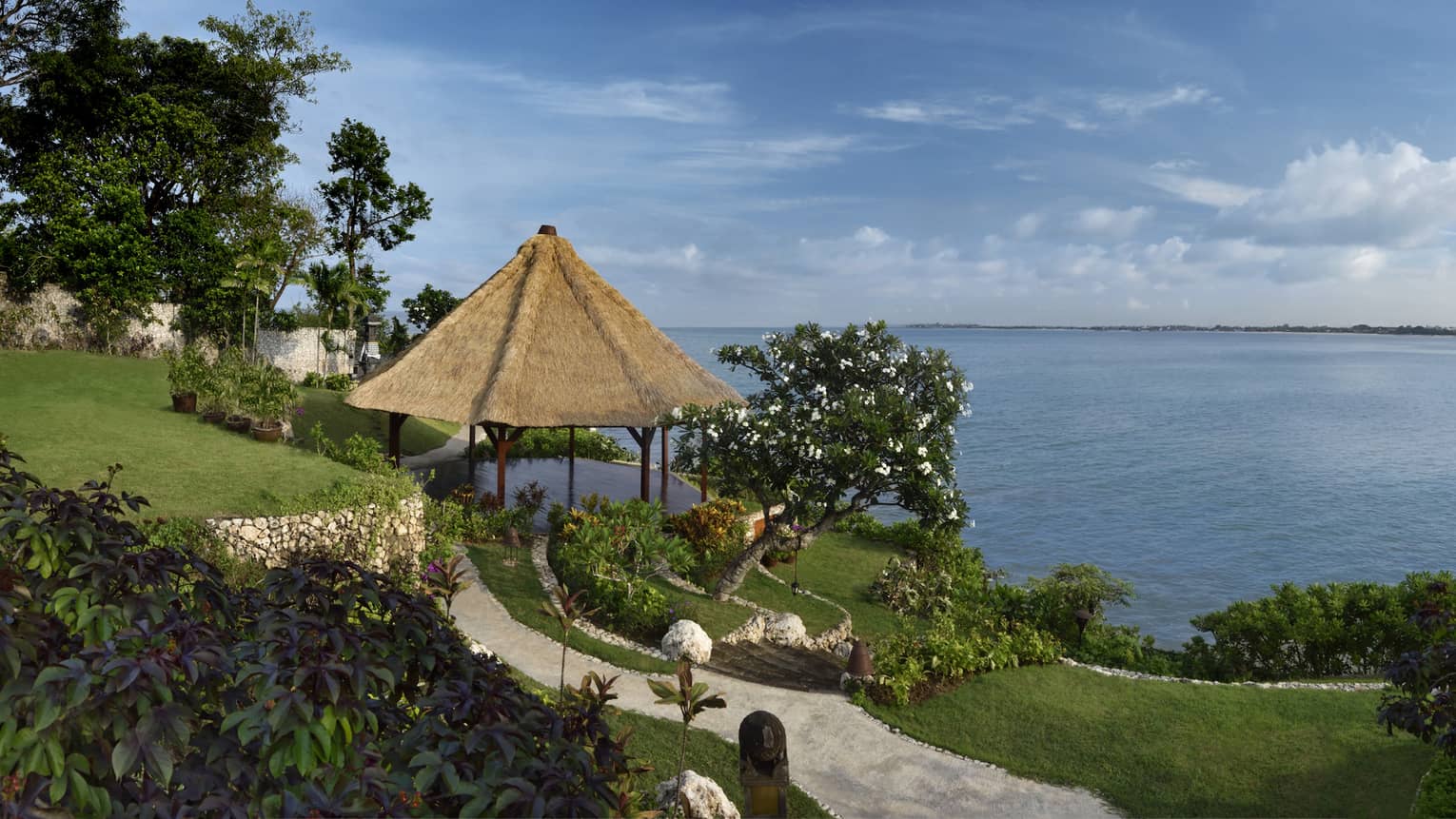 A paved trail runs through a tiered grassy area, past lush trees and a thatched hut bordering calm water under a cloudy sky.