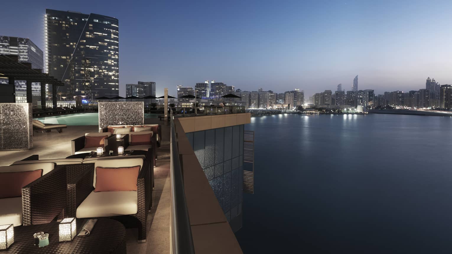 Hotel rooftop patio overlooking Arabian Gulf at night with wicker chairs with cushions, swimming pool