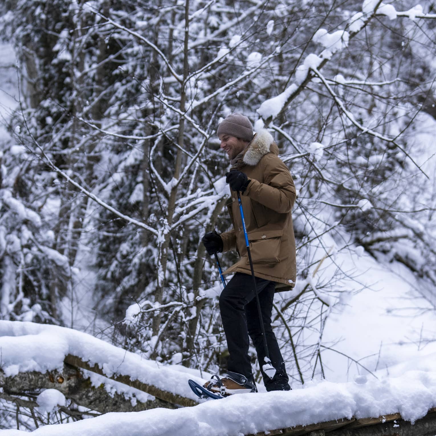 A snowshoer in a knit cap, parka and gloves uses poles to cross fallen logs in a dense, snow-covered forest.