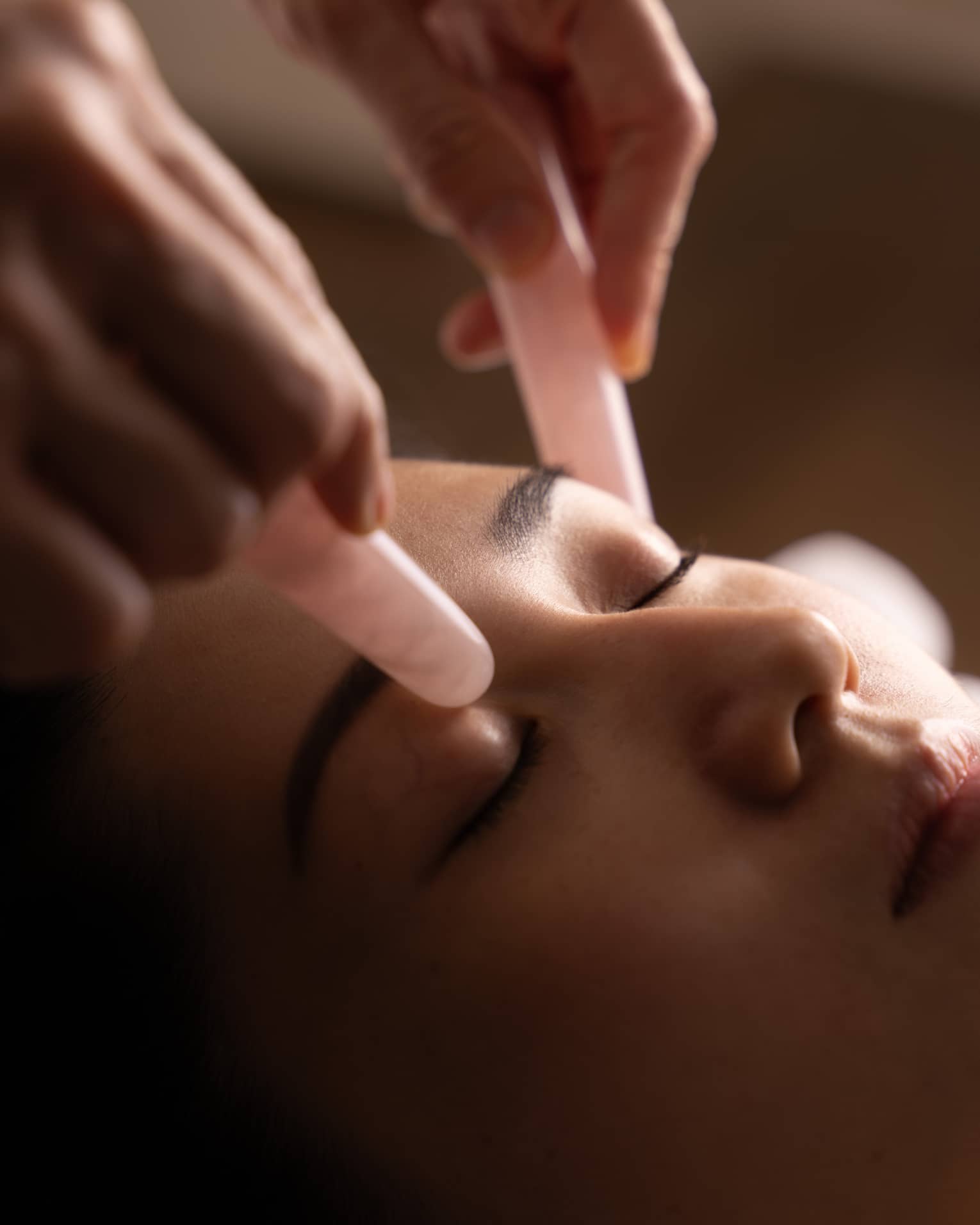 Hands hold pair of pink jade stones against woman’s eyelids during facial
