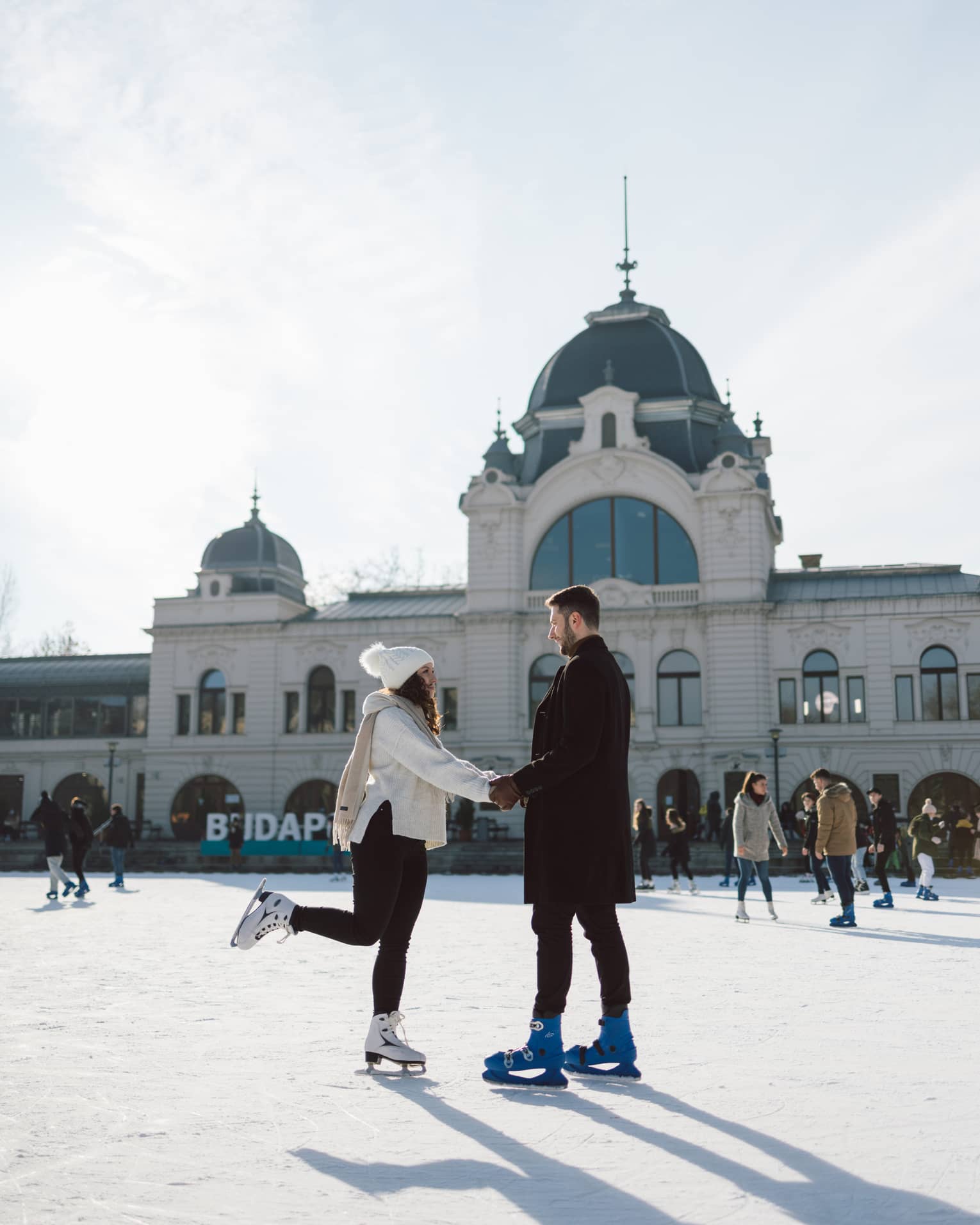 Couple dressed in warm clothing, holding hands while ice-skating on city rink