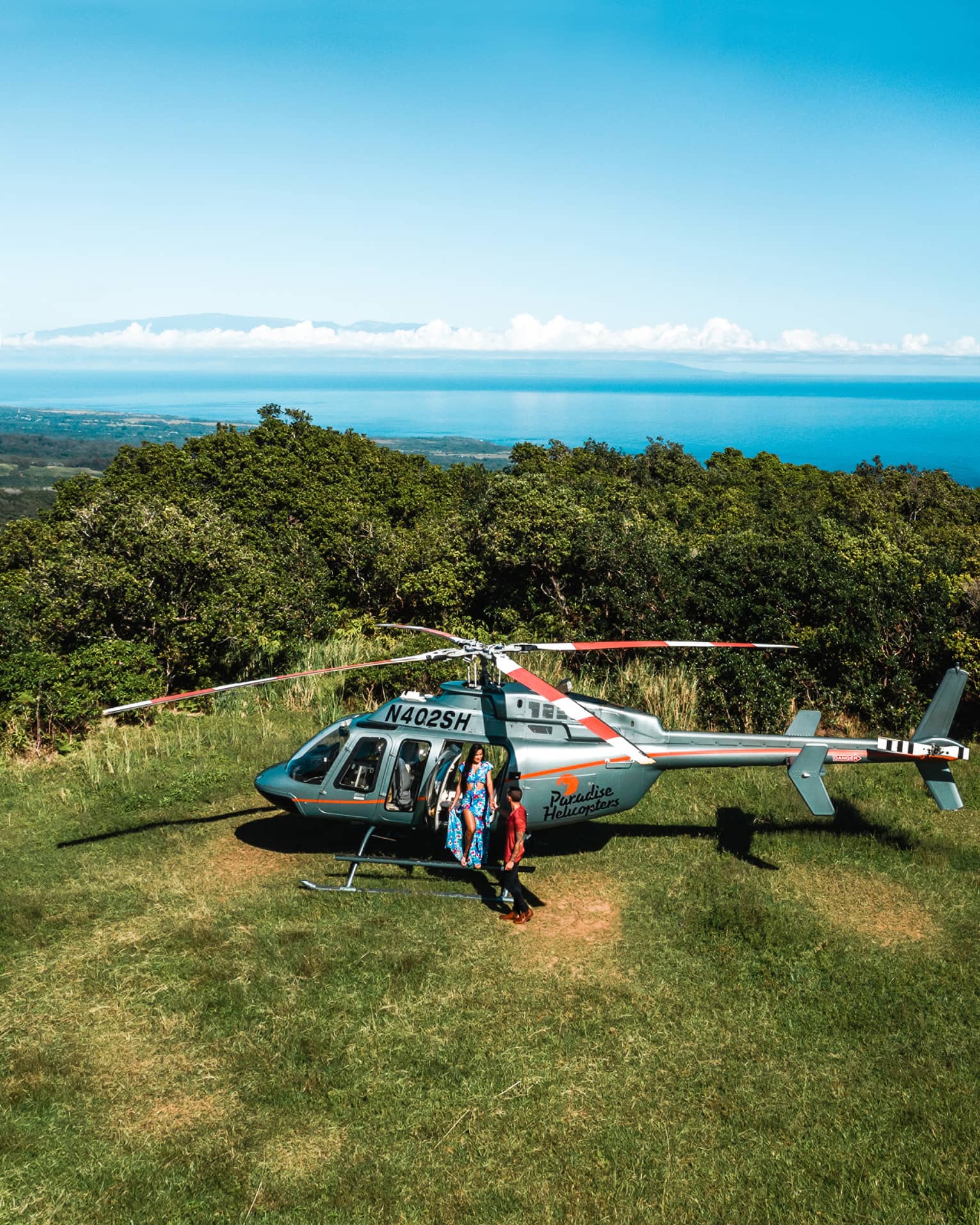 Woman and boy standing beside helicopter on grass with bushes and ocean water behind them