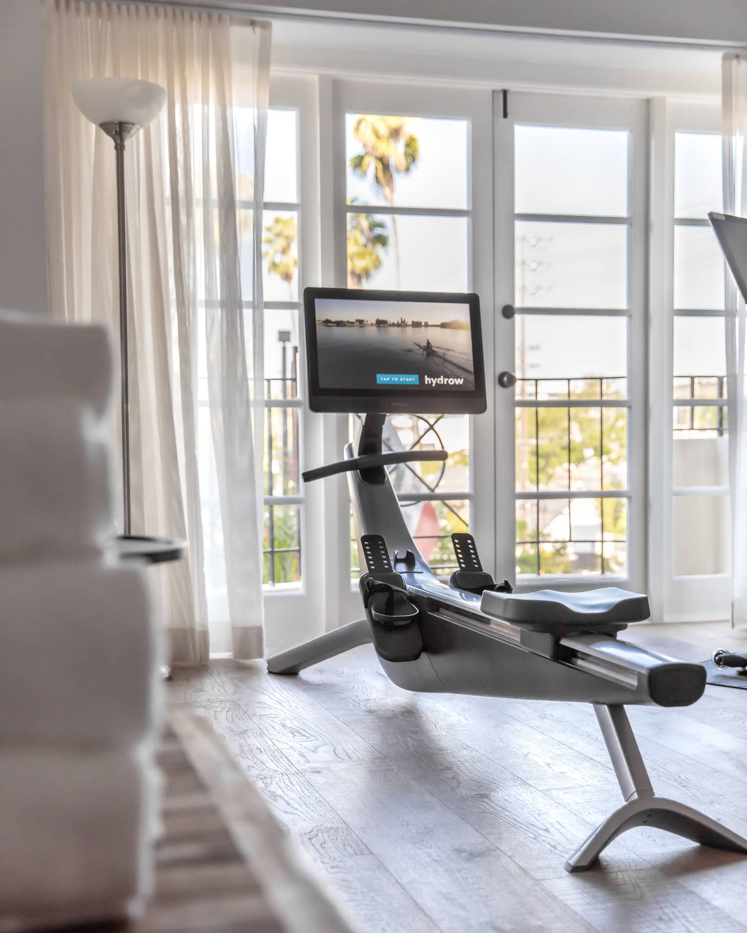 In a bright room, a rowing machine equipped with a screen faces two French doors, sheer white curtains pulled to the side.