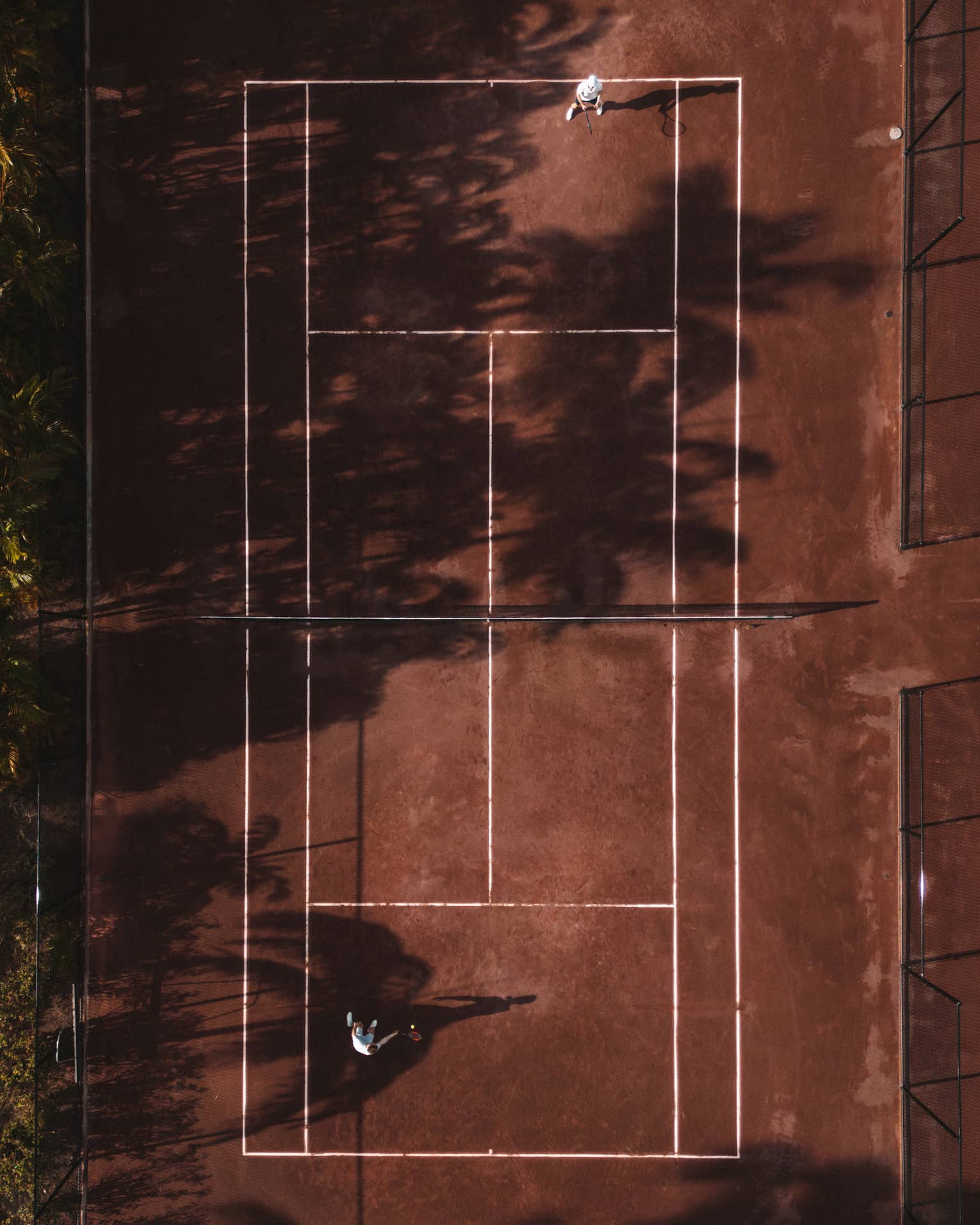 An aerial view of a red clay tennis court partially shaded by trees