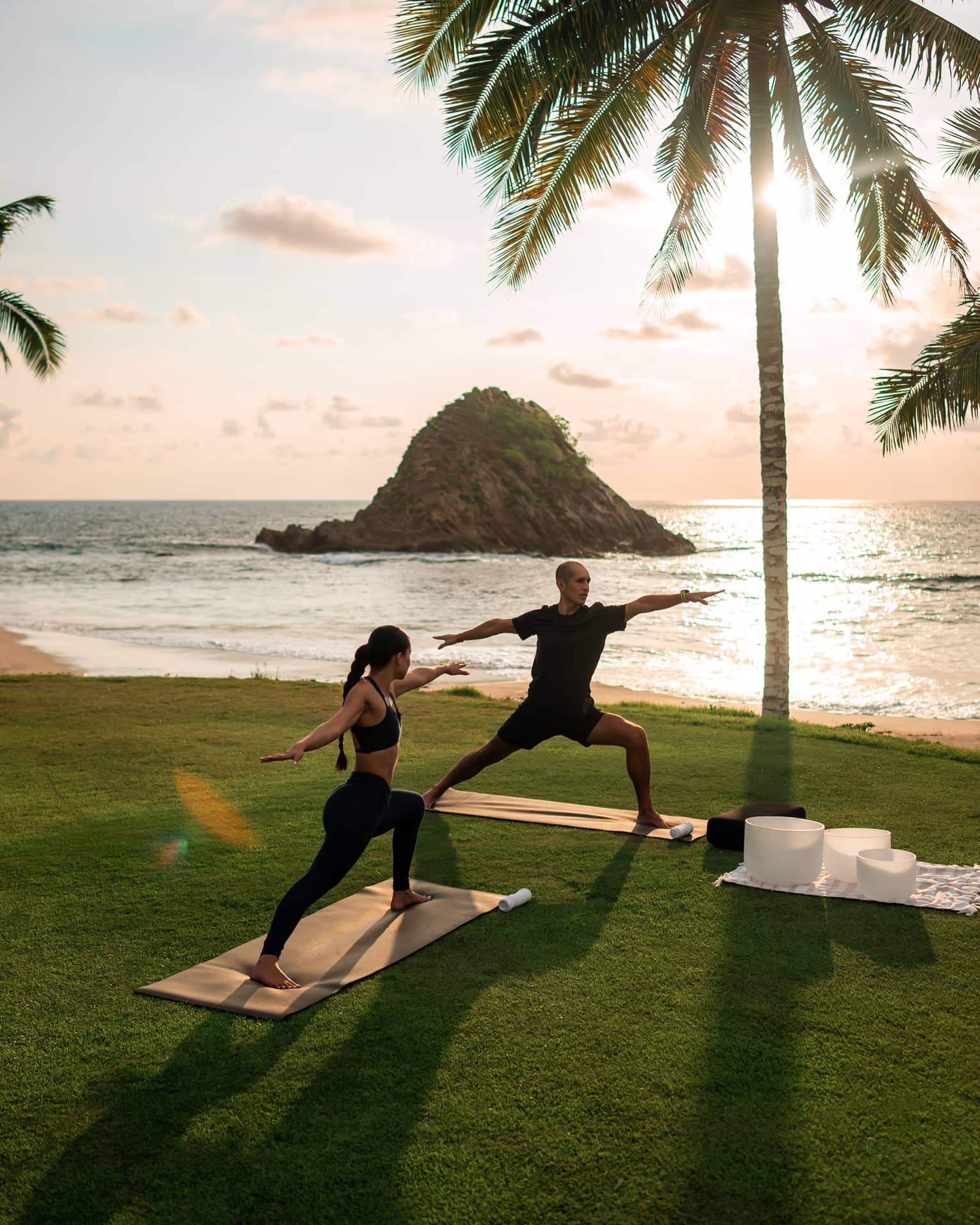 Two people in yoga poses on yoga mats on a lush grassy area next to the beach. A palm tree, the beach, the ocean and a bright but partially cloudy sky are in the background.