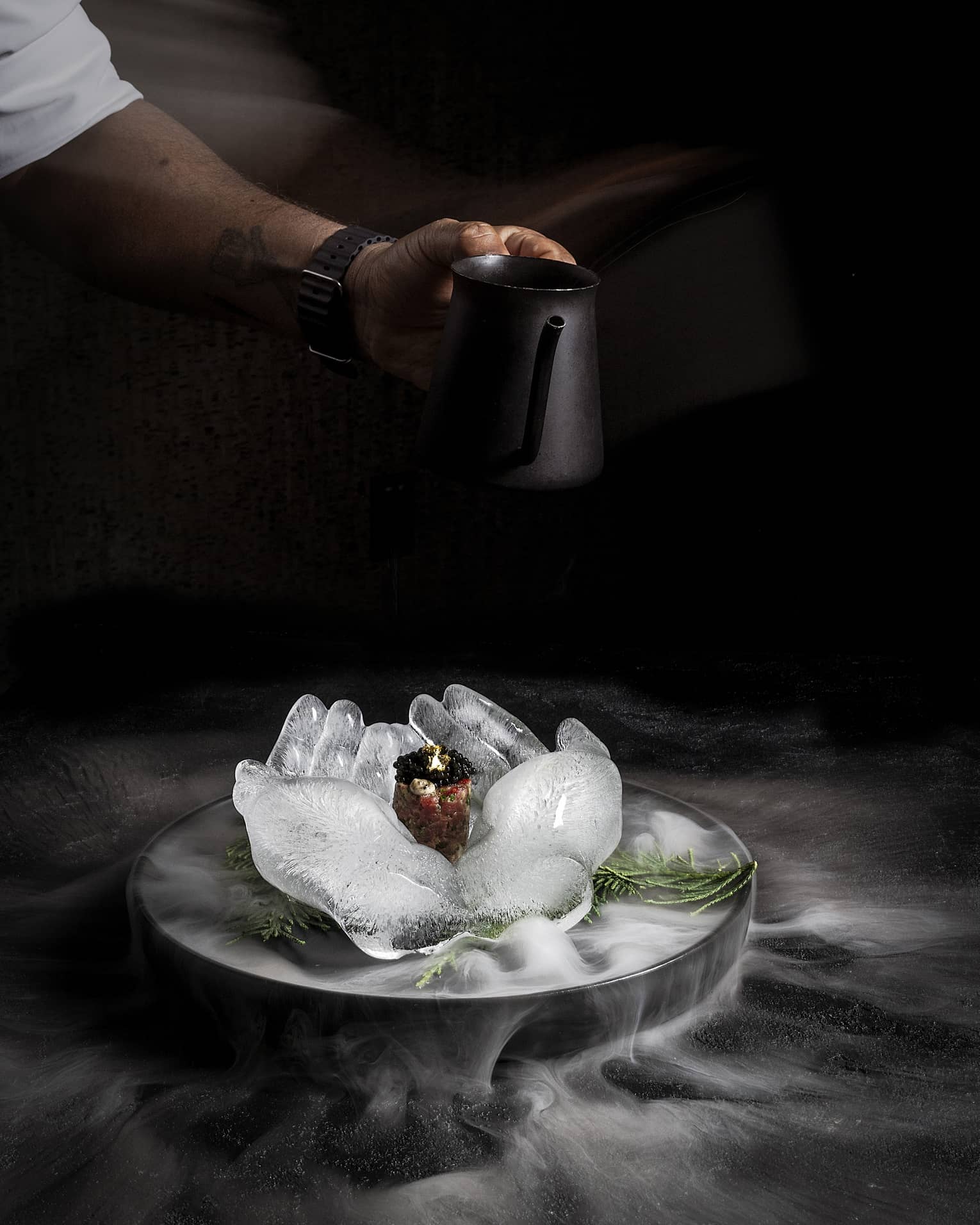 Food served on an ice sculpture shaped like two open hands with steam from dry ice floating around the black background