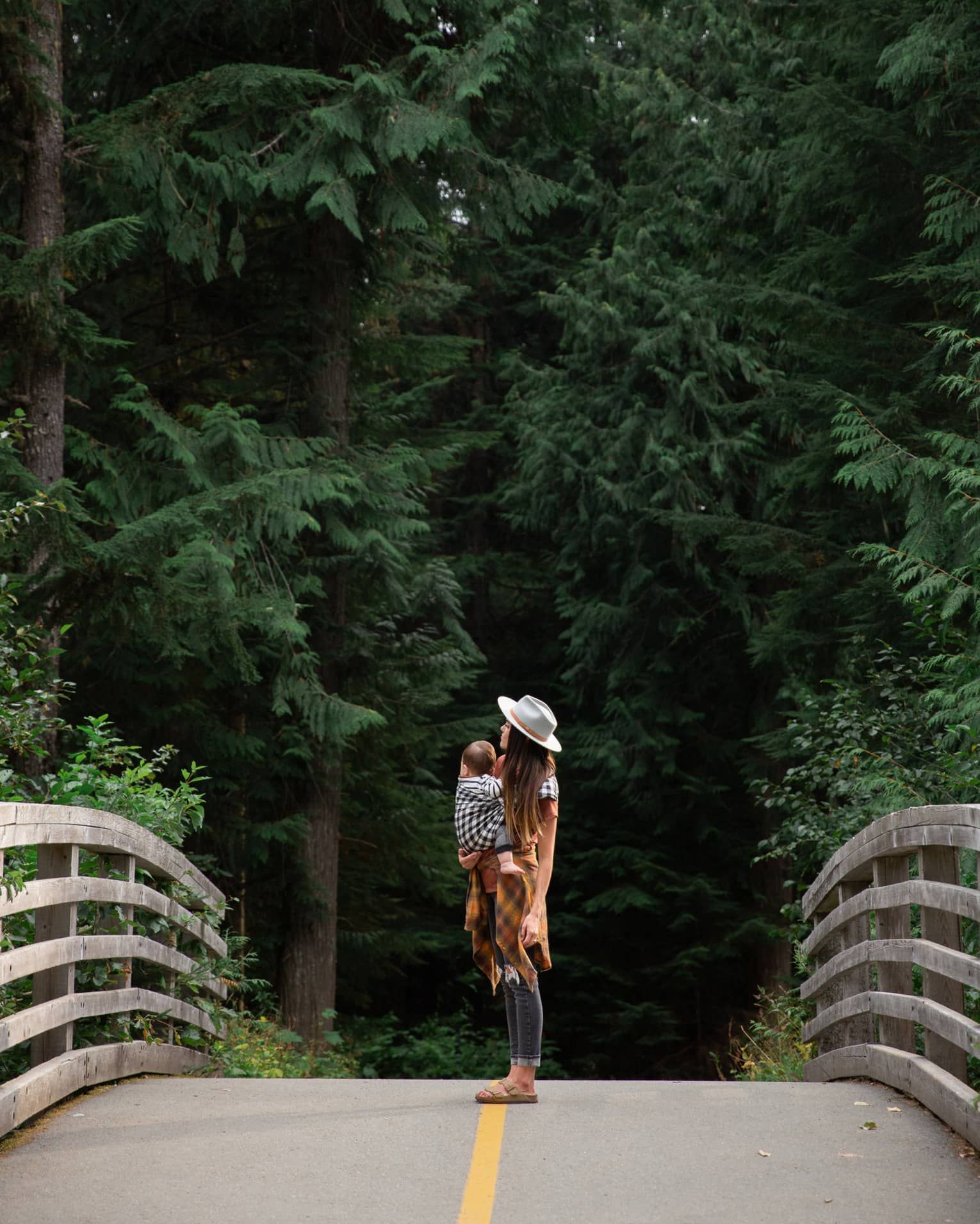 Woman with long dark hair wearing a beige hat, cognac-coloured long sweater and jeans holds a baby in a sling and stands in the middle of the bridge with tall green trees in the background