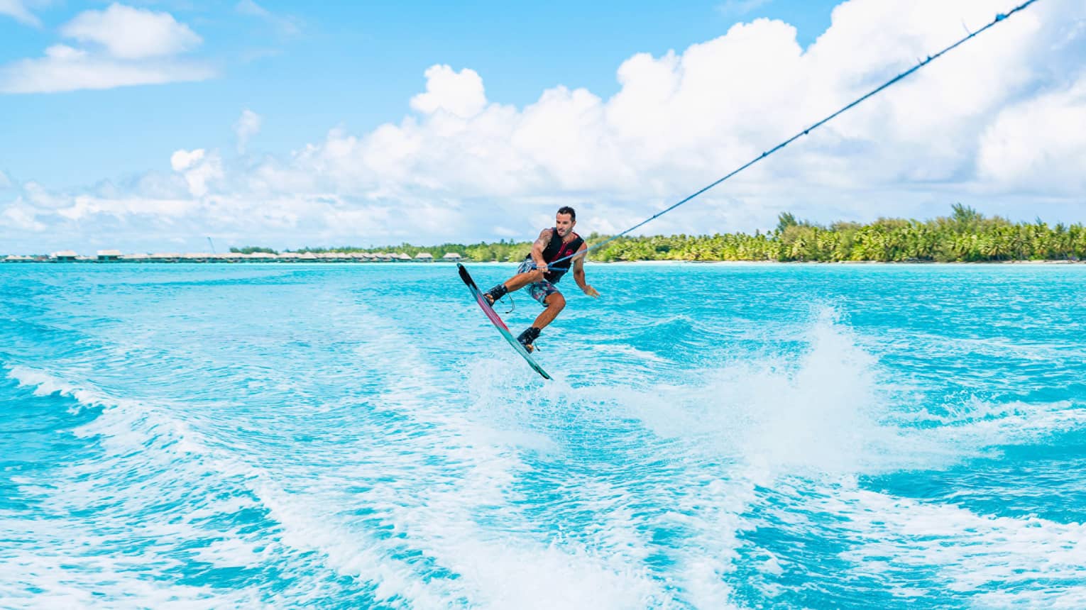A wakeboarder leaps above the waves, one hand holding a taut rope as they're towed through the ocean beneath a sunny sky.