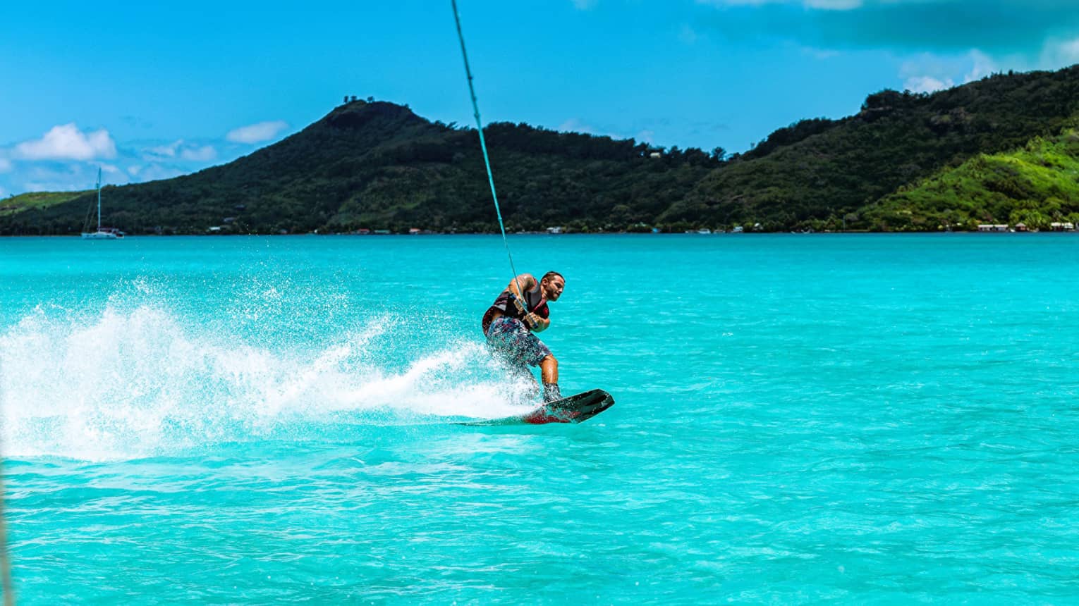 A guest creates a spray of water with their wakeboard as they're towed through the ocean before a backdrop of forested hills.
