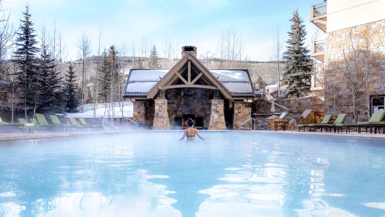 A woman in a pool with a cabin in front of her and snow covered trees around her.