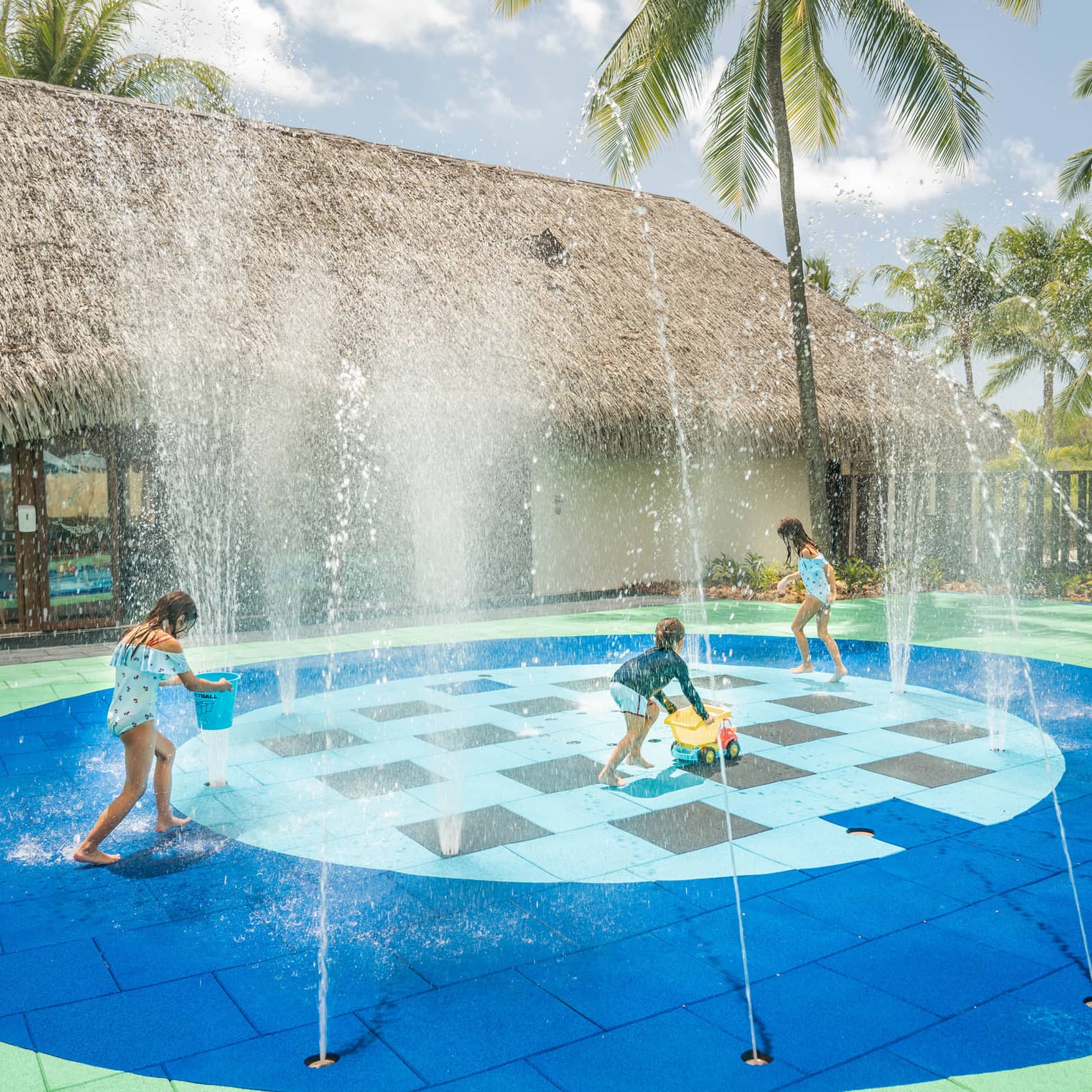 Group of children in swimsuits play on splash pad next to grass-roof building