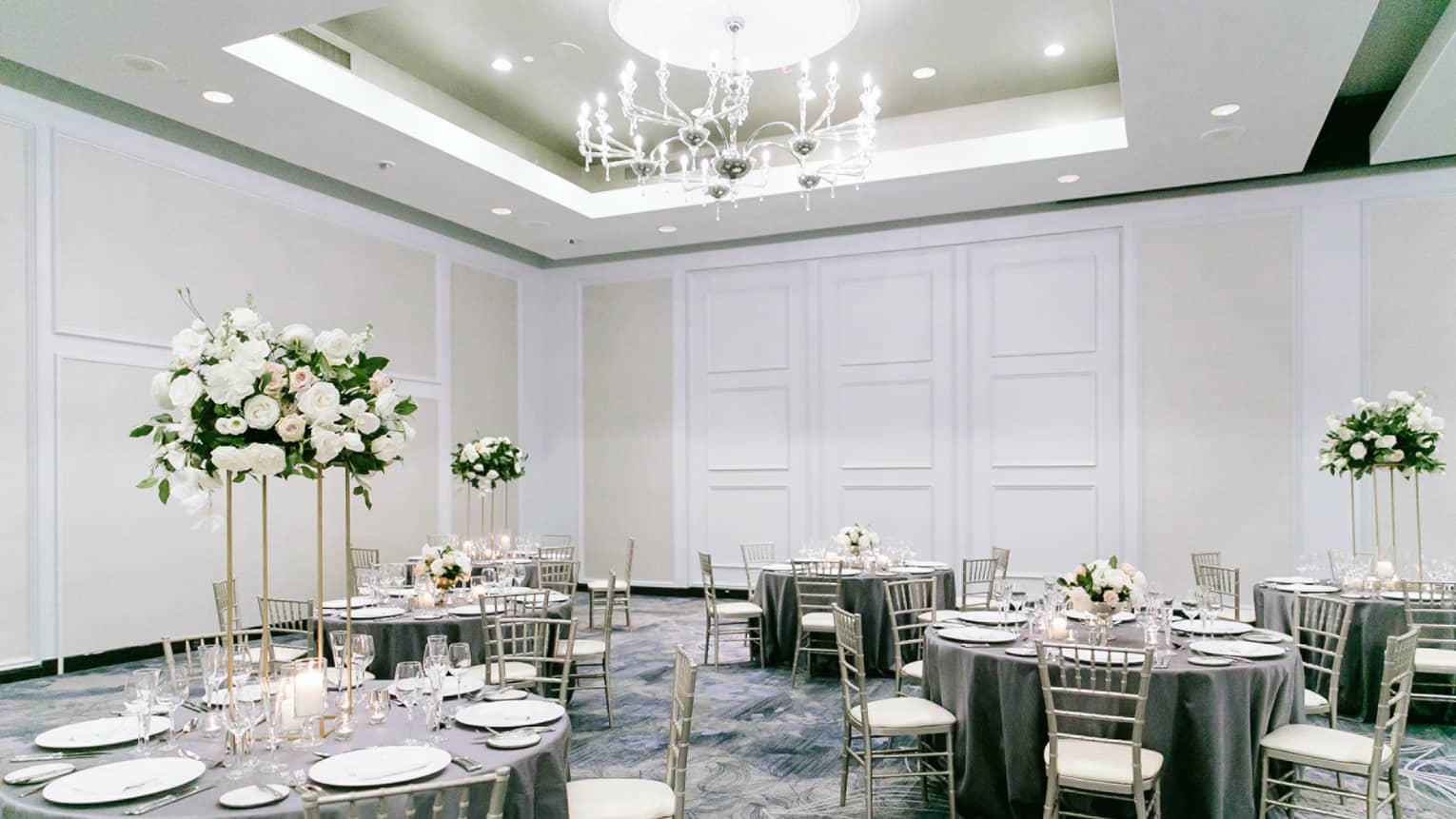 A reception area with grey clothes round tables and white wooden chairs, flower centerpieces, and a large chandalier.