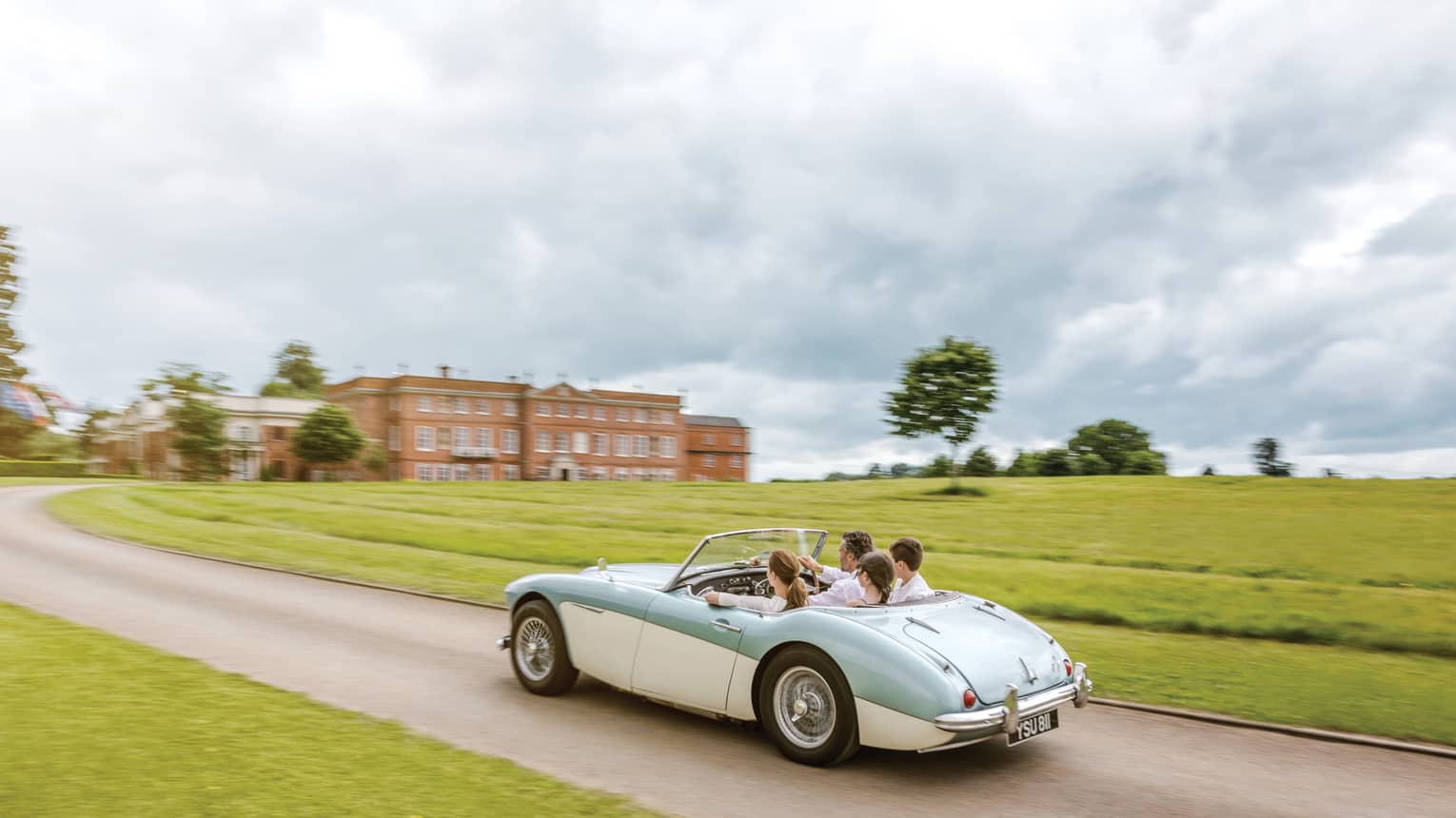 Family rides in vintage convertible car down road past English countryside to hotel manor