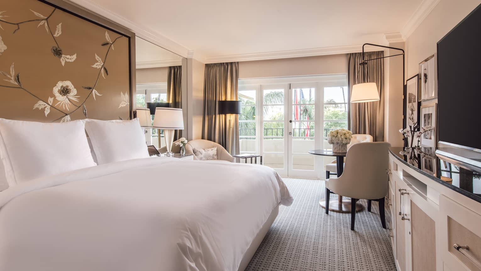 Deluxe Balcony Room hotel bed against tall floral headboard across from TV, dressers, chairs by balcony
