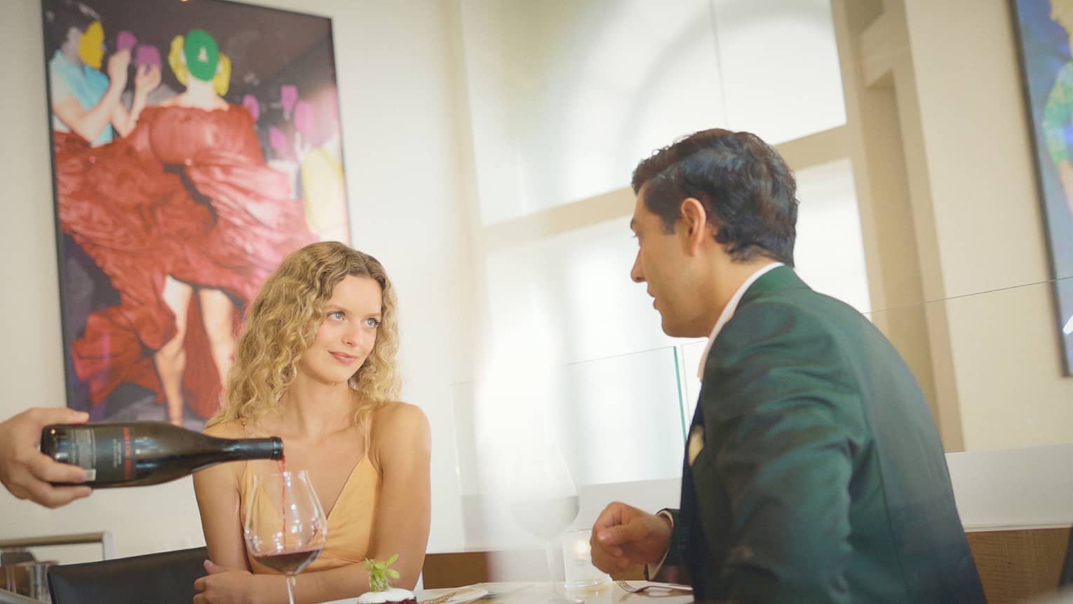A couple sits across from each other in a restaurant dining room as a server pours wine into a glass on the table